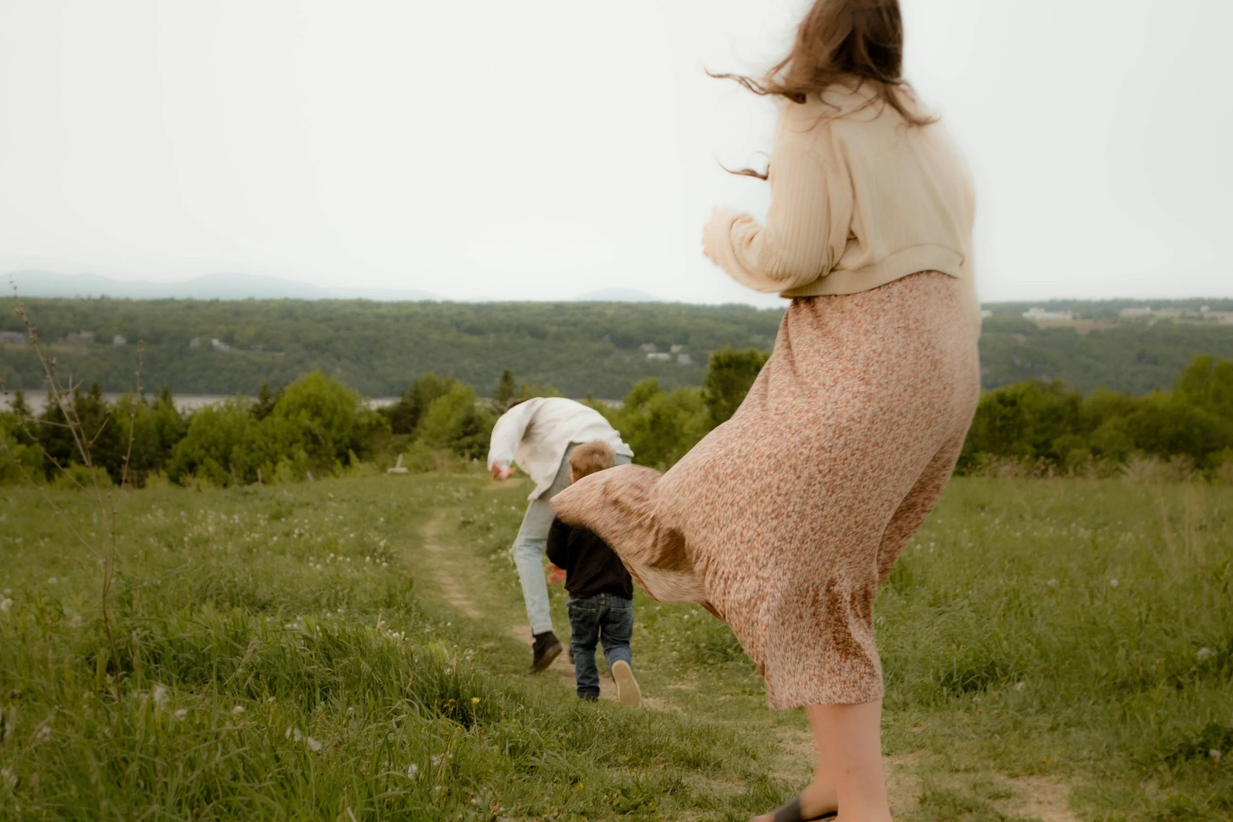 Une femme marche dans un champ avec deux enfants, un chien blanc et un paysage de collines en arrière-plan.