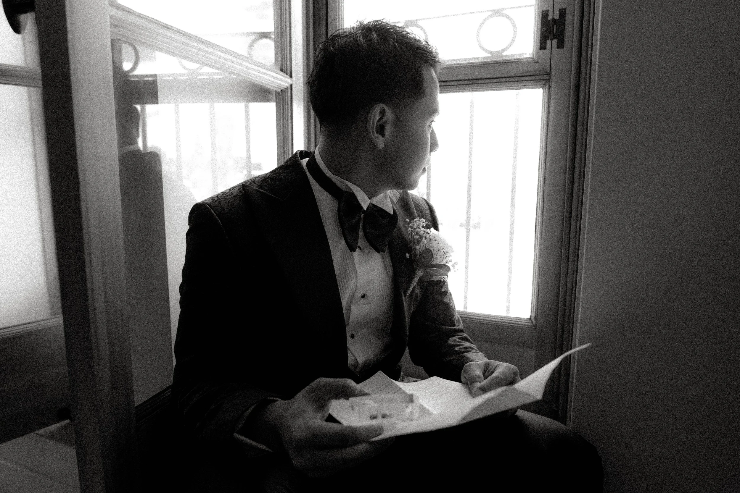 Un homme regarde par la fenêtre tout en lisant un document, habillé en costume avec une décoration boutonnière, dans une pièce éclairée par la lumière naturelle.