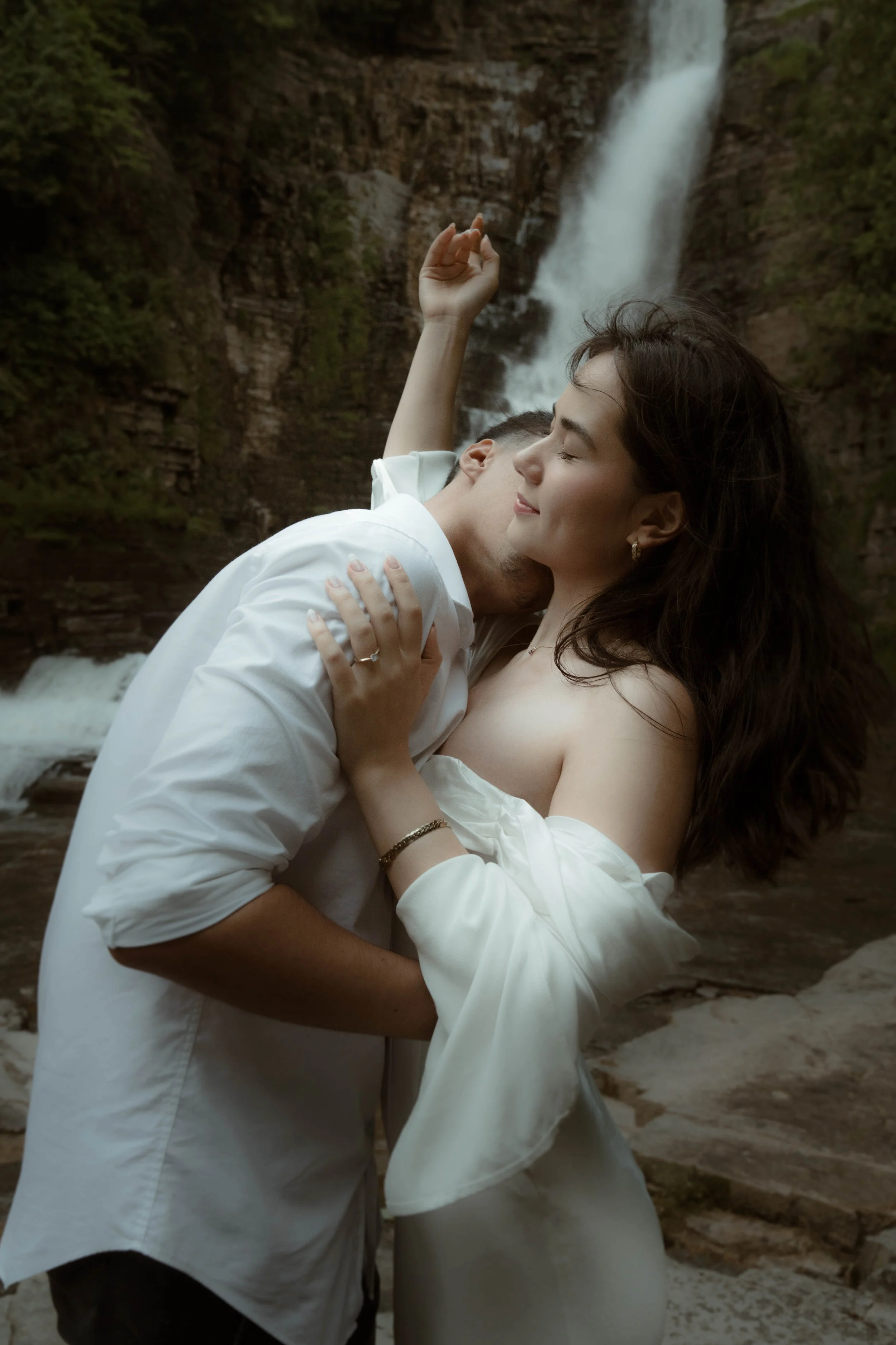 Un couple en vêtement blanc s'embrasse près d'une cascade dans un environnement naturel.
