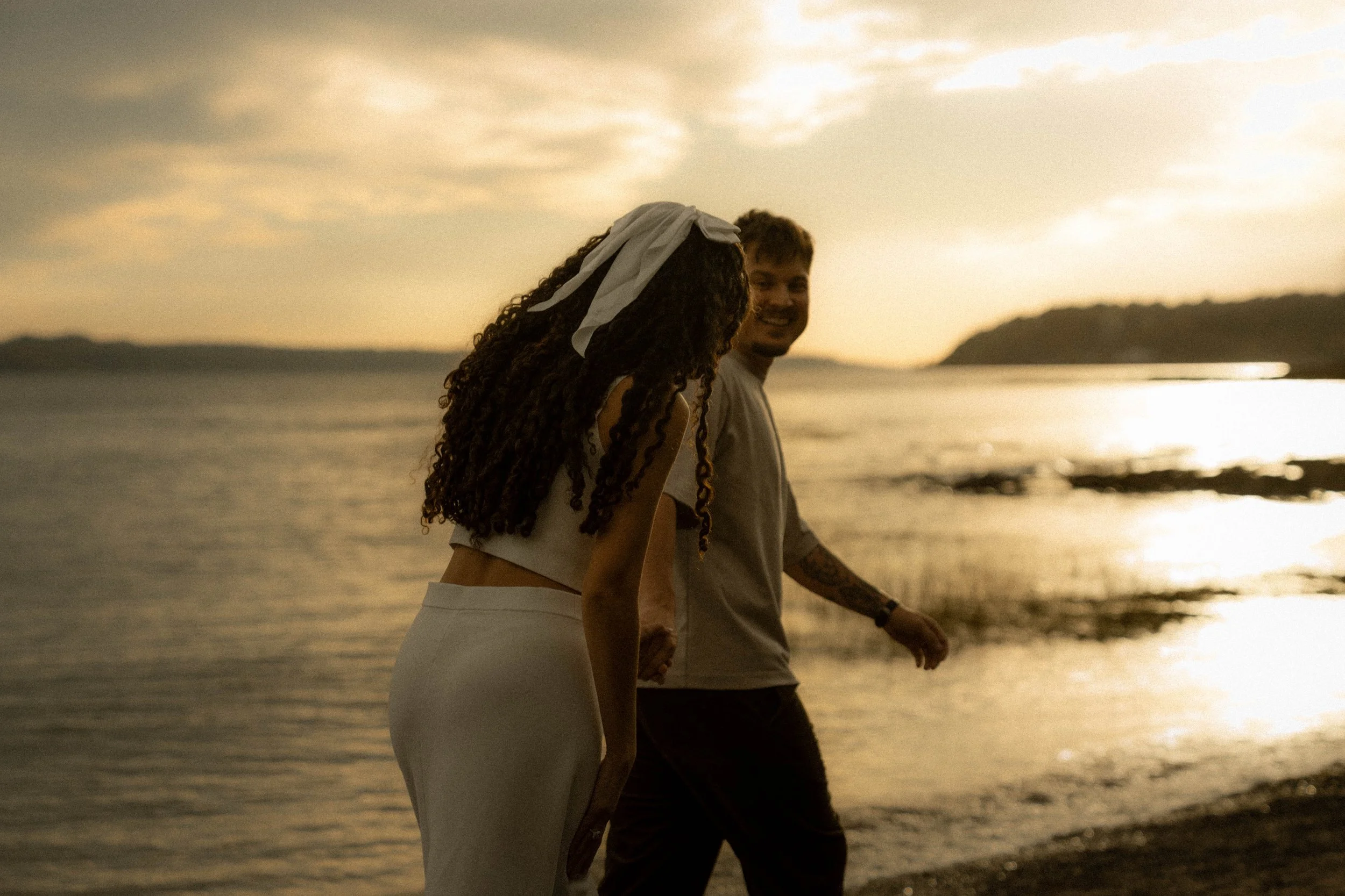 Un couple marche sur la plage au coucher du soleil, la femme porte un bandeau blanc dans ses cheveux bouclés et un haut blanc, l'homme porte un t-shirt gris clair.