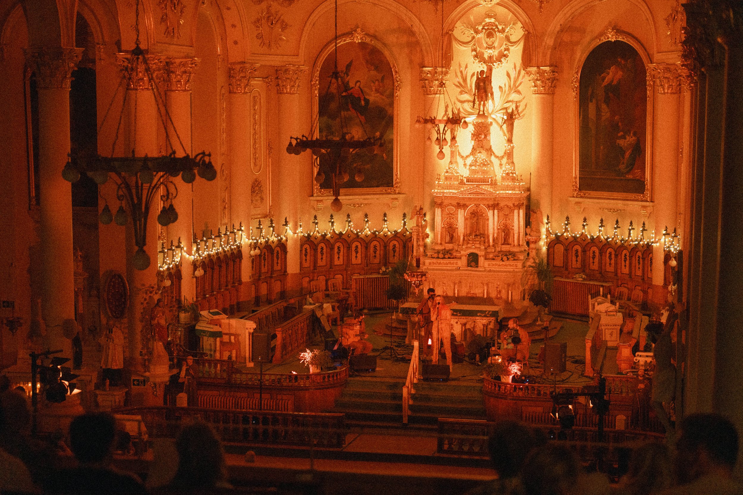 Intérieur d'une église ou d'une cathédrale ornée de fresques, chandeliers et guirlandes lumineuses, avec des personnes assises ou se tenant debout.