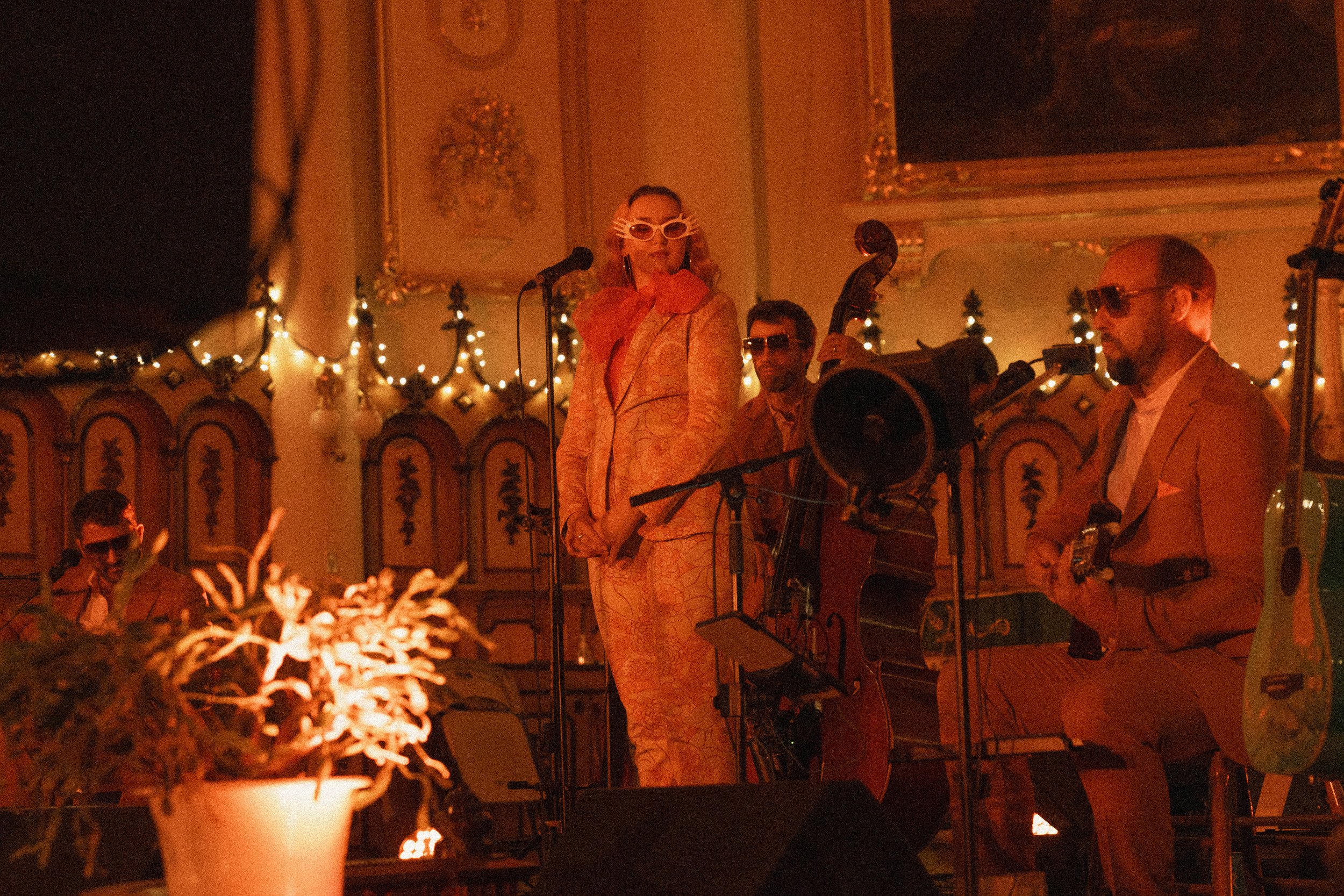 Une scène de concert ou de performance artistique dans une salle décorée avec des guirlandes lumineuses, où trois musiciens jouent avec un éclairage chaud. La femme au centre porte des lunettes blanches, une robe élégante couleur pêche et une écharpe