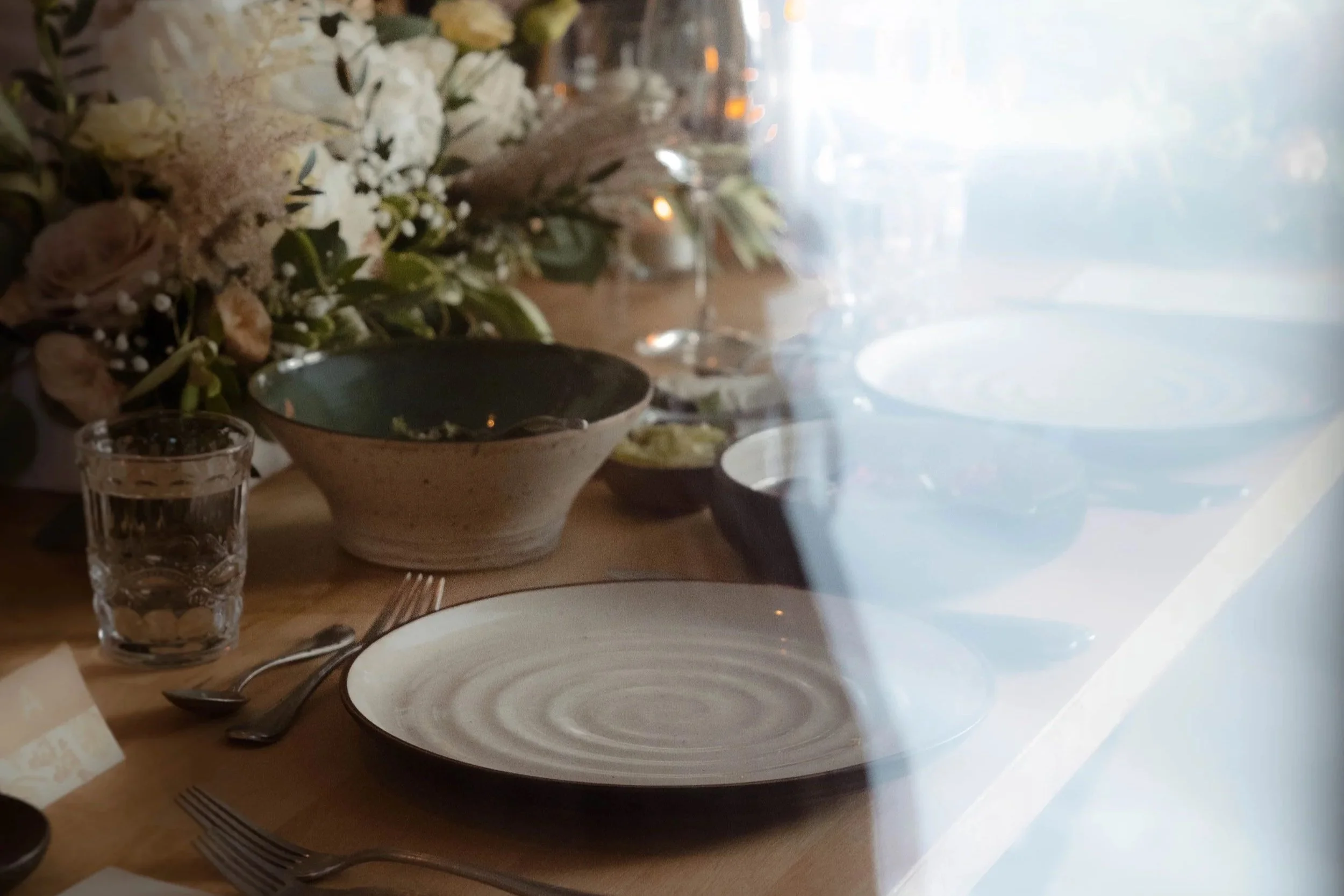 Une table prête pour un repas, avec des assiettes, des couverts, un verre d'eau, des bols, des fleurs en décoration et une fenêtre derrière avec une lumière naturelle