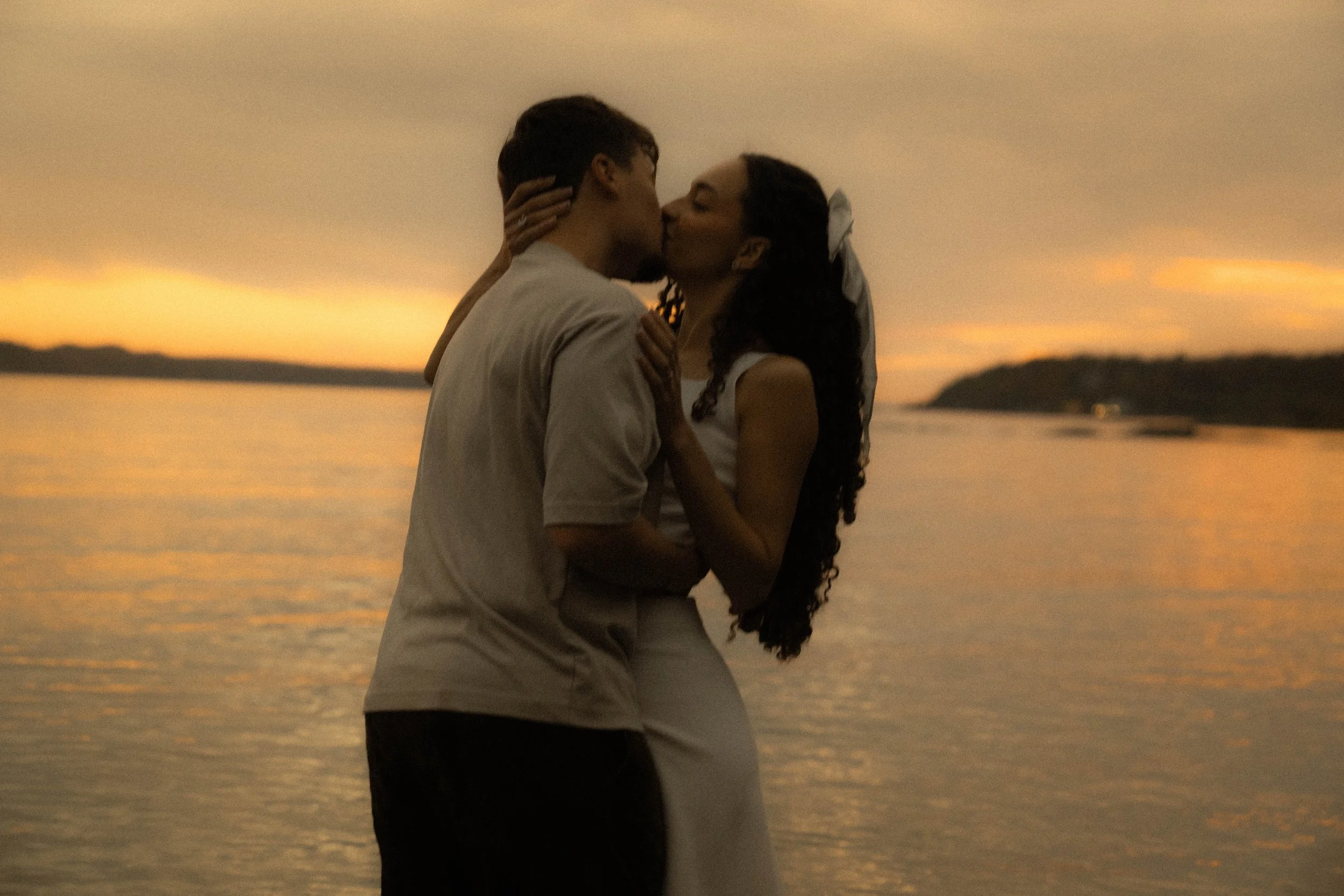 Une jeune femme et un jeune homme s'embrassent près d'un lac au coucher du soleil, dans un paysage naturel avec des collines à l'horizon.