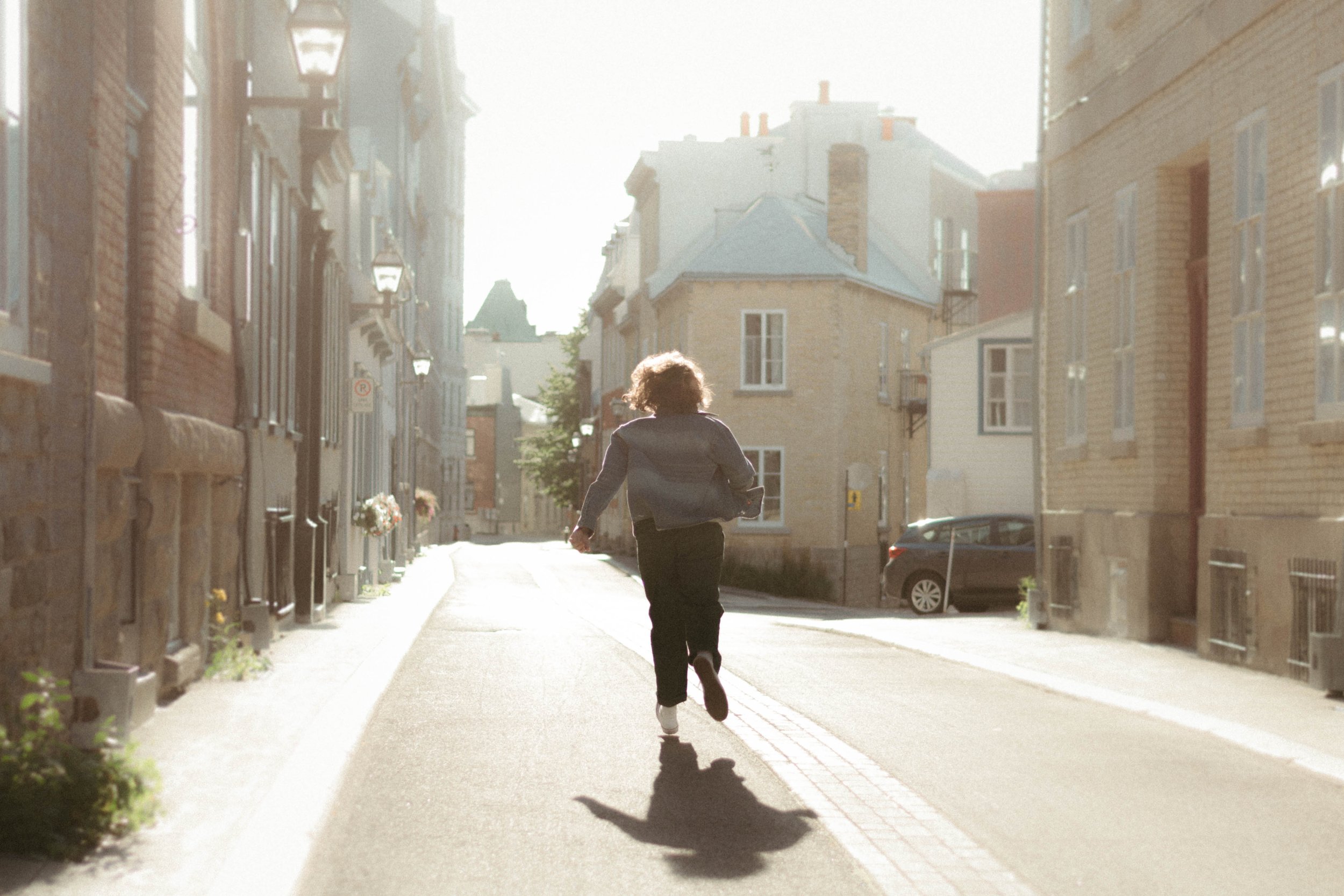Une personne marche dans une rue pavée en plein jour, entourée d'immeubles en briques et de lampadaires, avec un soleil brillant derrière les bâtiments.