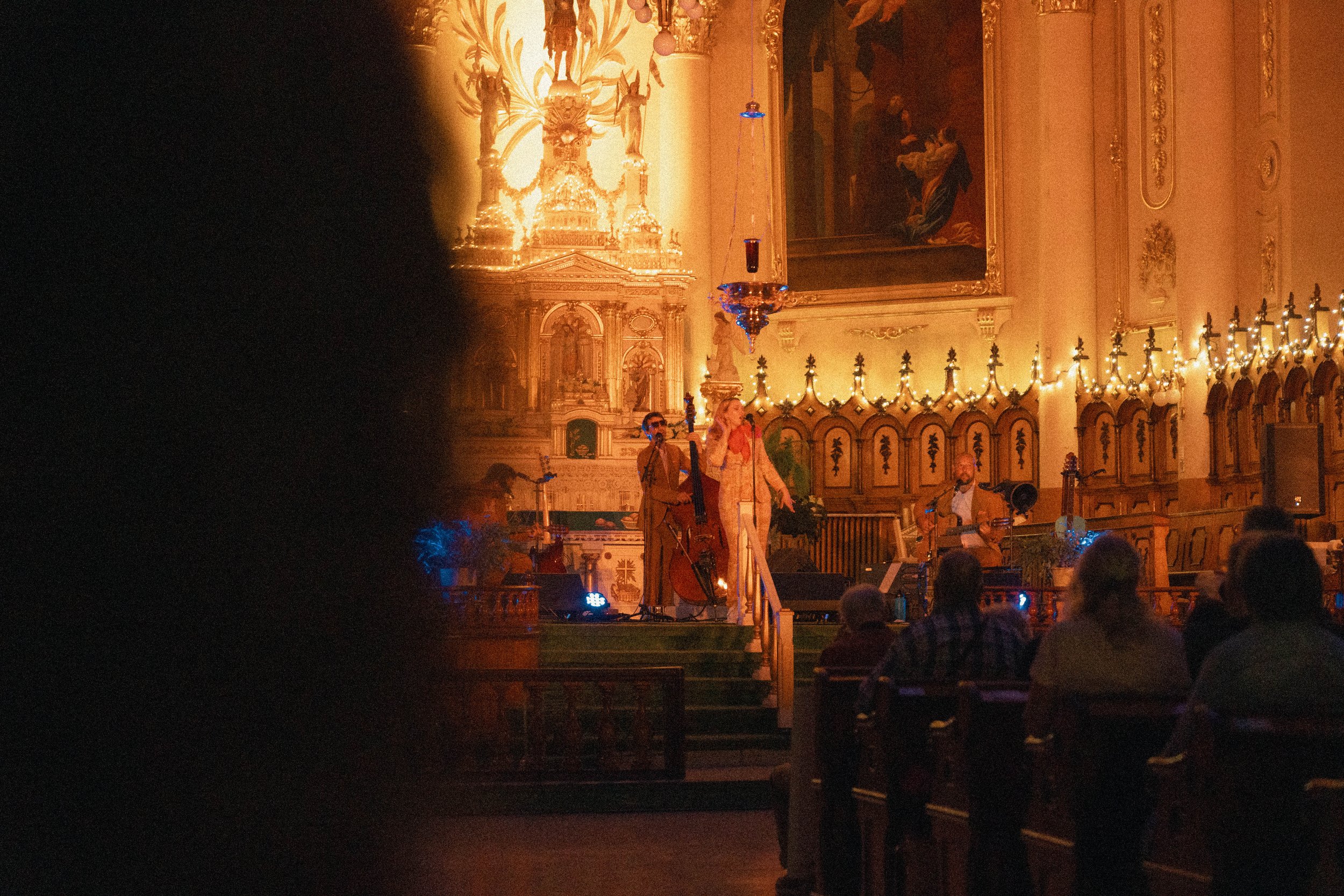 Une scène musicale dans une salle ornée de dorures, avec un groupe de musiciens jouant sur scène et des personnes assises dans l'assistance.