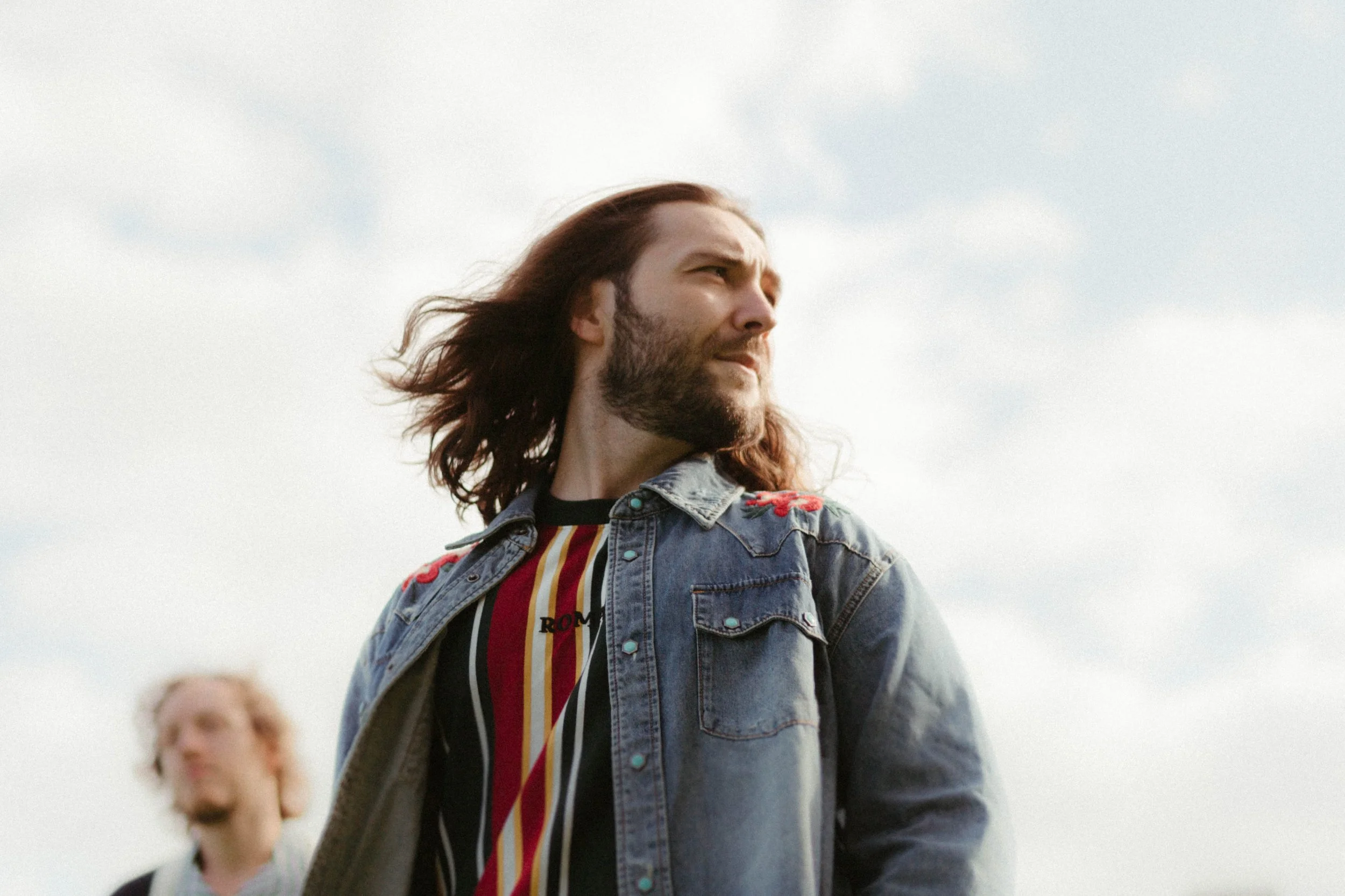 Un homme avec des cheveux longs, barbe, portant une veste en jean et un t-shirt rayé, regarde au loin. Il y a un autre personne floue en arrière-plan. Le ciel est clair et dégagé.