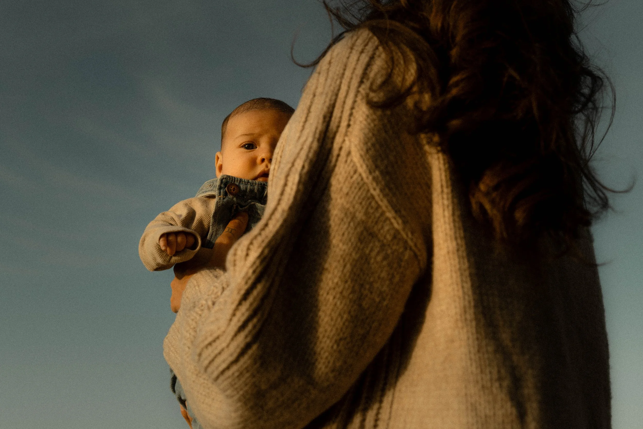 Une femme porte un bébé qui regarde vers l'objectif, avec un ciel clair en arrière-plan.