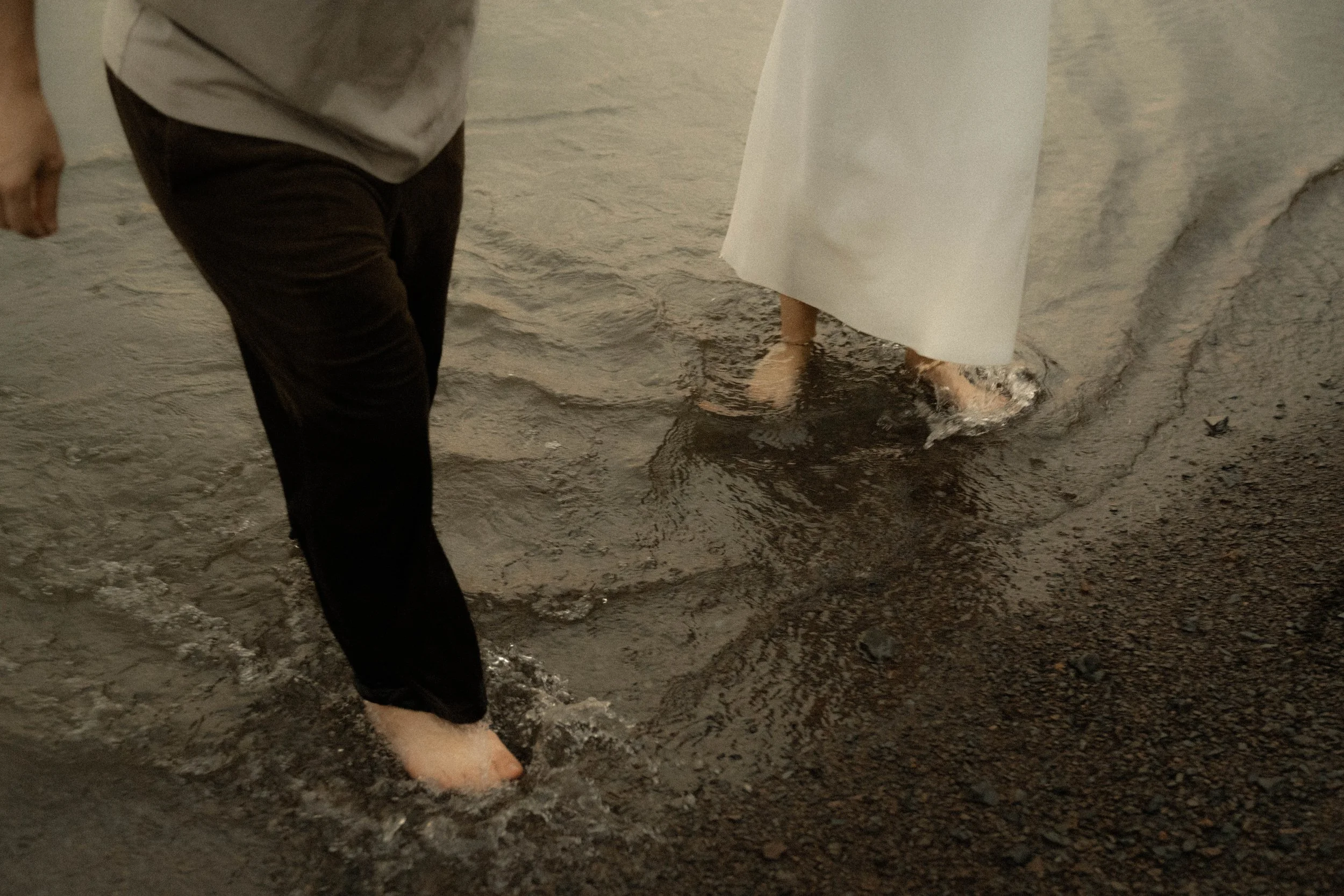 Personne marchant dans l'eau peu profonde, portant une longue robe blanche, pieds nus, sur une plage rocheuse.