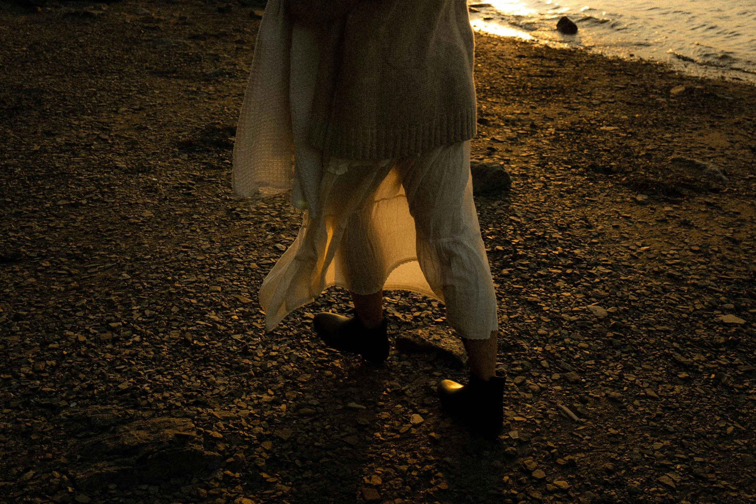 Personne marchant sur une plage rocheuse au coucher du soleil, portant des vêtements amples et des bottes