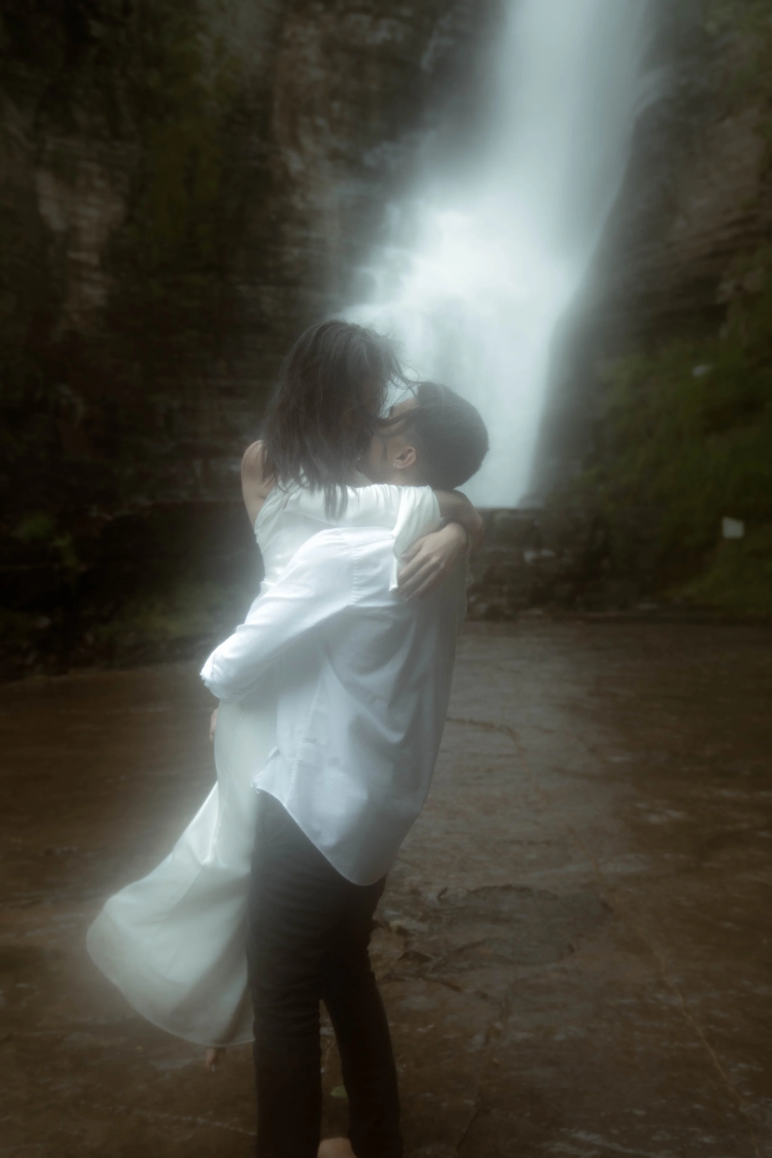 Deux personnes se tiennent sous une cascade dans un environnement naturel, l'une porte un chemisier blanc et l'autre des vêtements gris foncé.