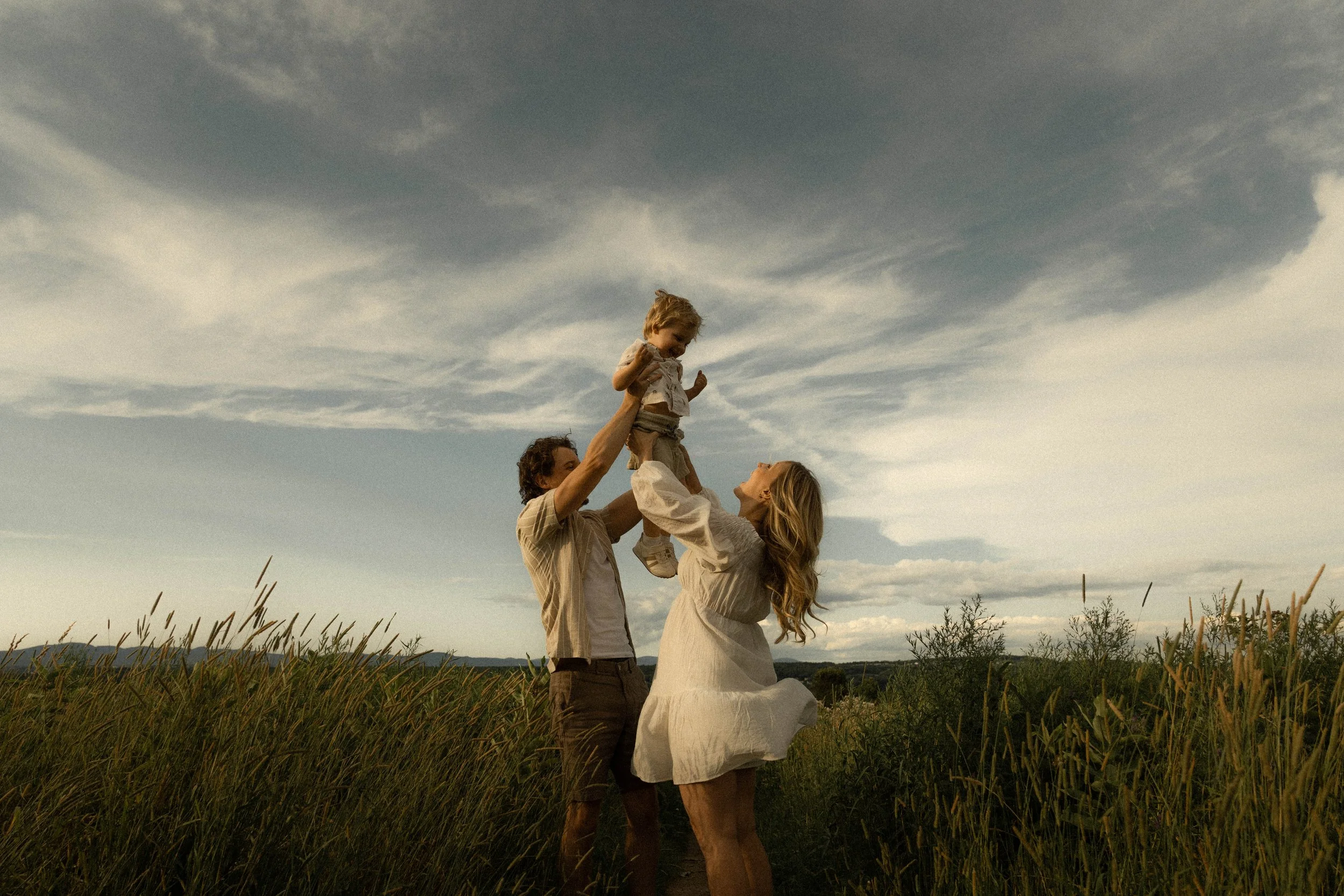 Un couple avec deux enfants dans un champ, l'un des enfants est soulevé en l'air par le père tandis que la mère regarde, sous un ciel nuageux.