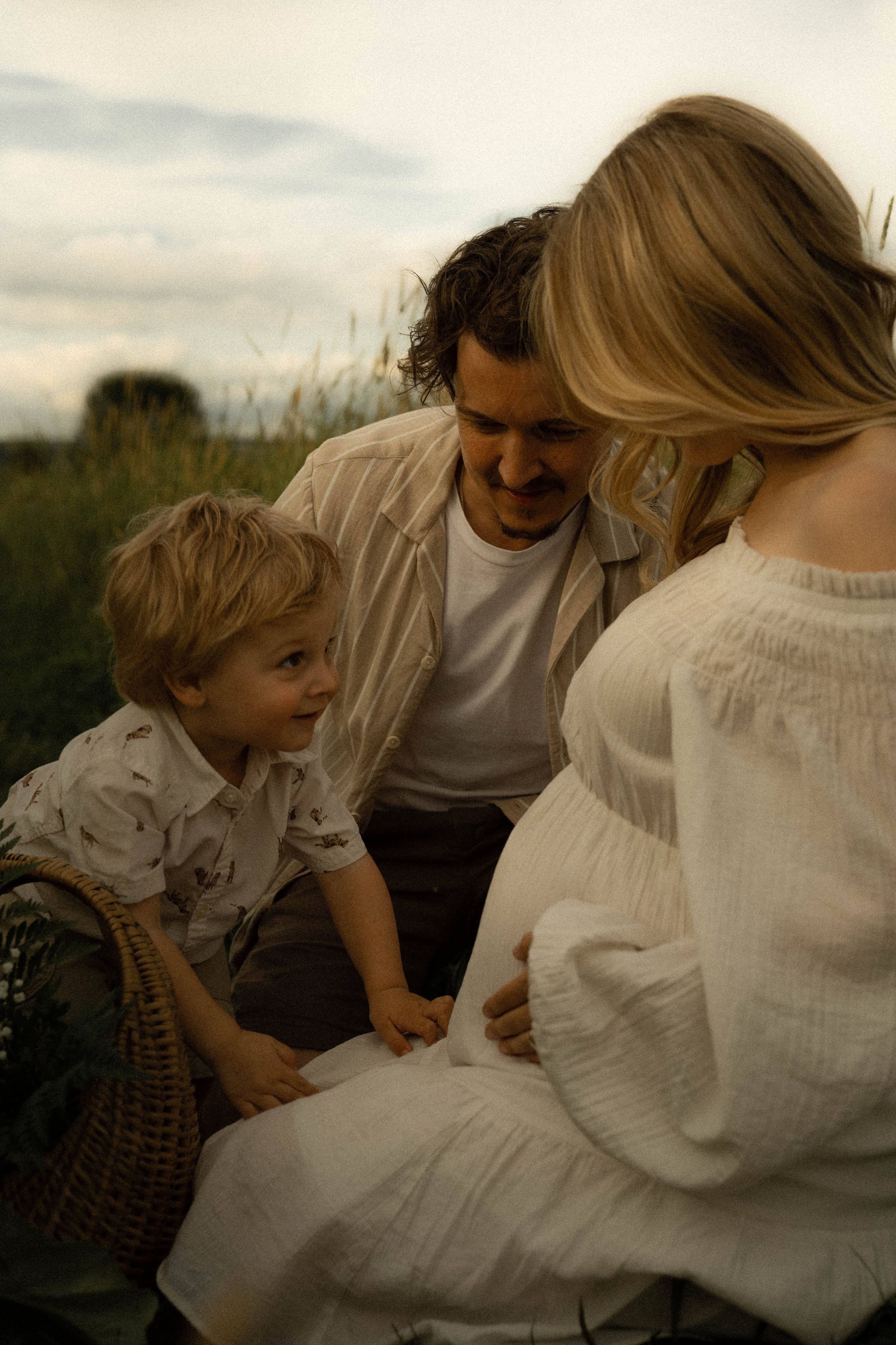 Une famille réunie dehors, une femme enceinte, un homme et un jeune garçon, tous souriants et se tenant la main.