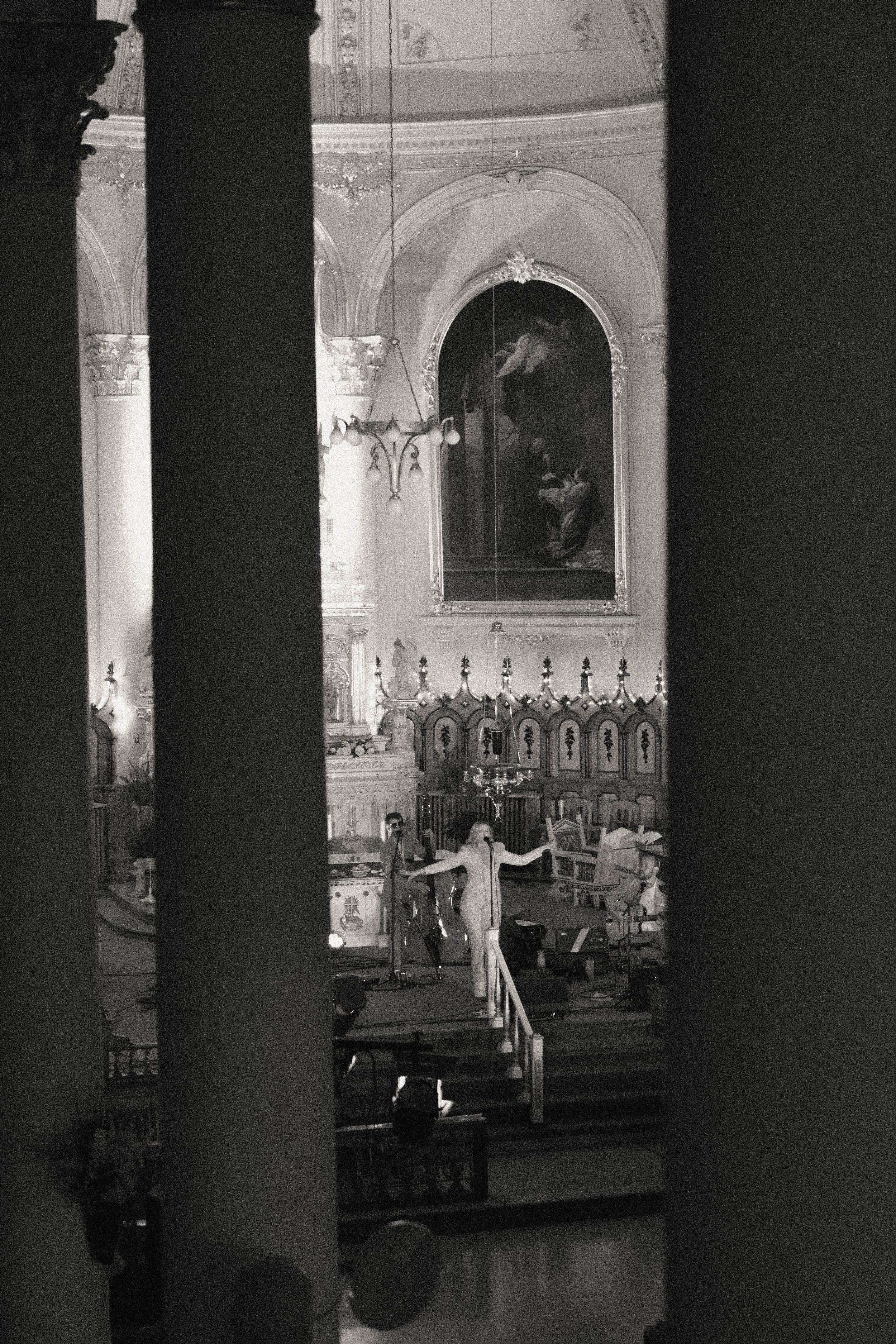 Une scène à l'intérieur d'une église ou d'une cathédrale avec des personnes en costume, probablement pour un concert ou une représentation, avec un décor religieux en arrière-plan, notamment une grande peinture ou icône, des colonnes ornées et une gr