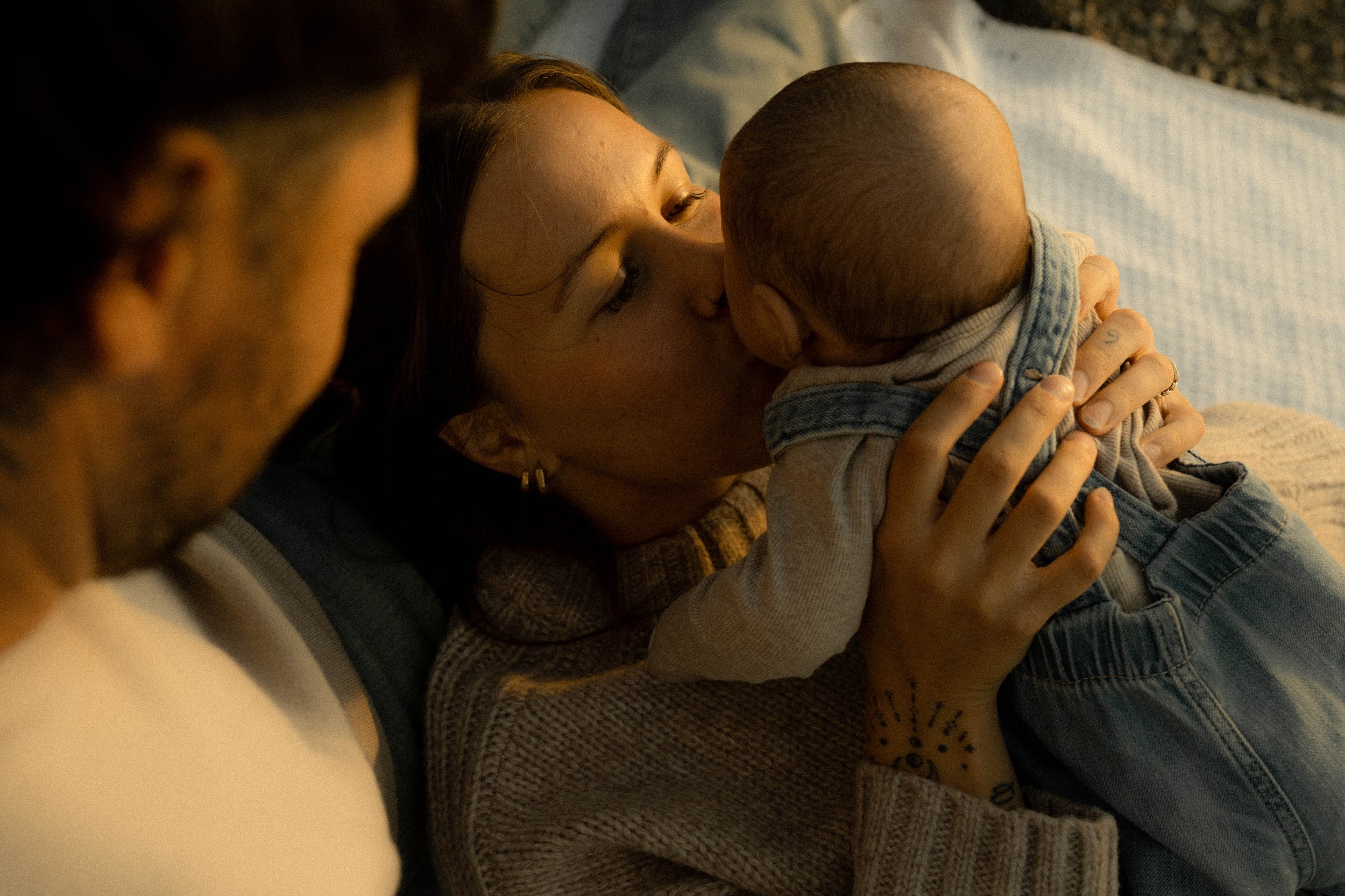 Une femme est en train d'allaiter un bébé. Autour d'elle, deux autres personnes sont présentes, une à gauche dont seul un fragment du visage est visible, et une autre à l'arrière, dont la tête est censée être présente mais est hors cadre.