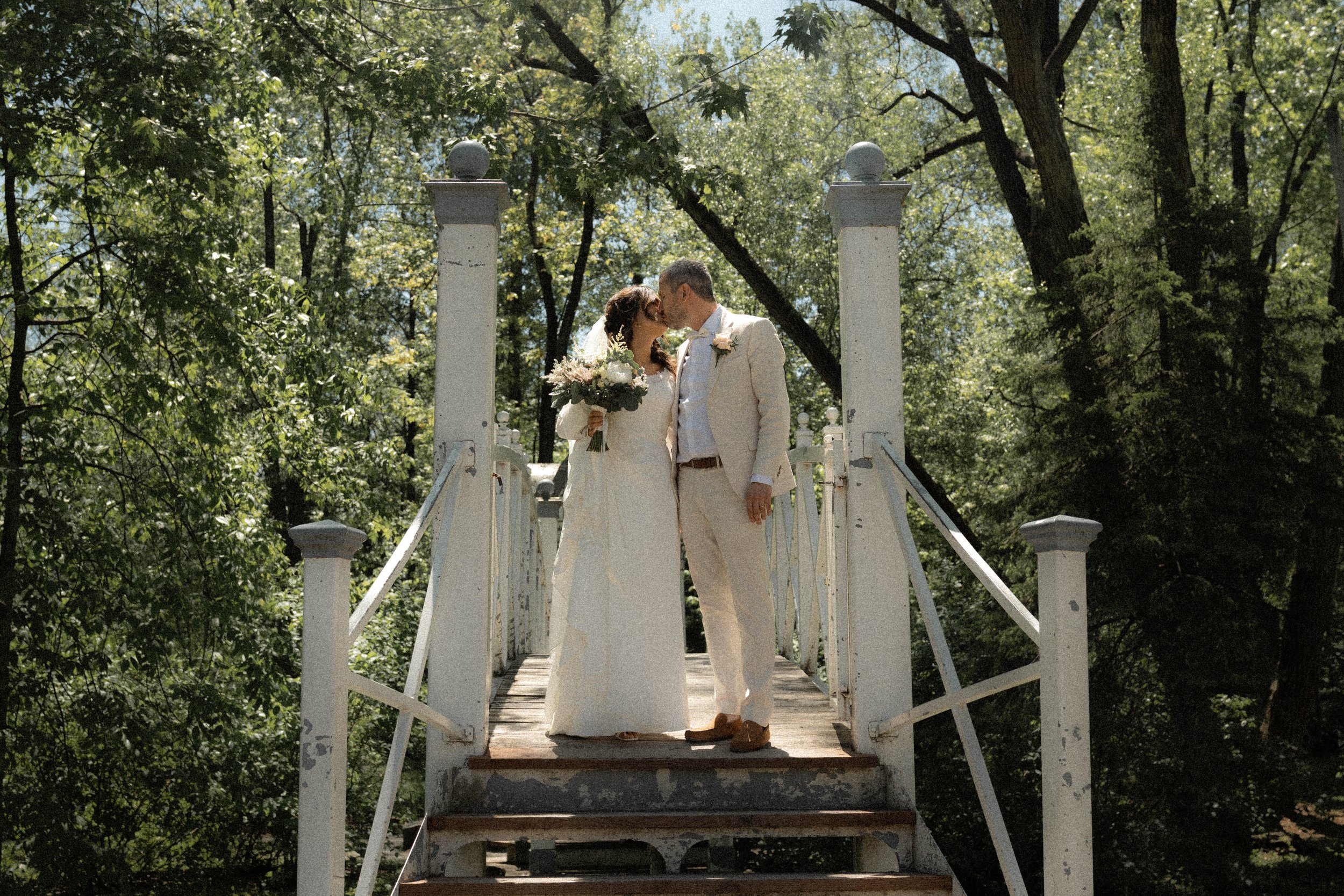 Un couple en mariage s'embrassant sur un pont en bois dans un parc entouré d'arbres.