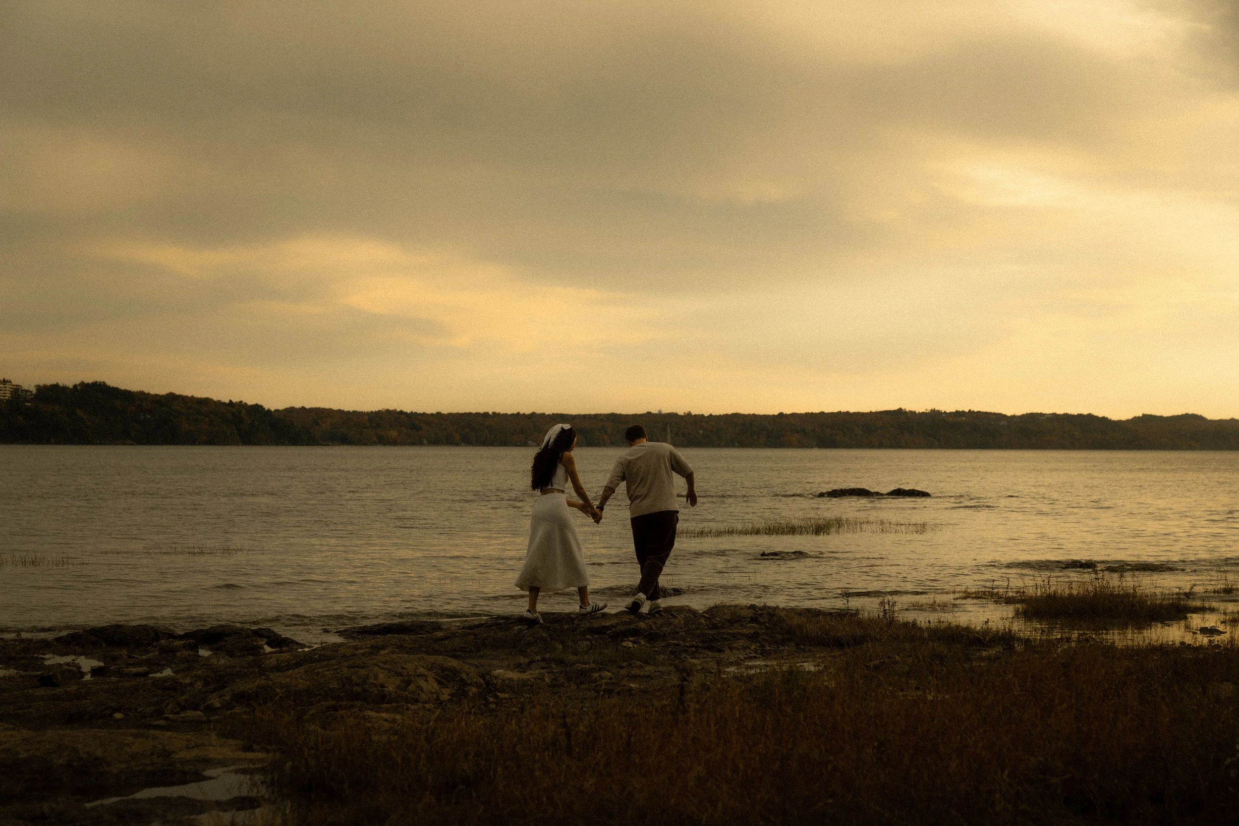 Un couple marche main dans la main au bord d'une rivière ou d'un lac au coucher du soleil, la femme porte une robe blanche ou claire et l'homme un t-shirt gris, horizon avec des collines ou forêt et ciel nuageux.