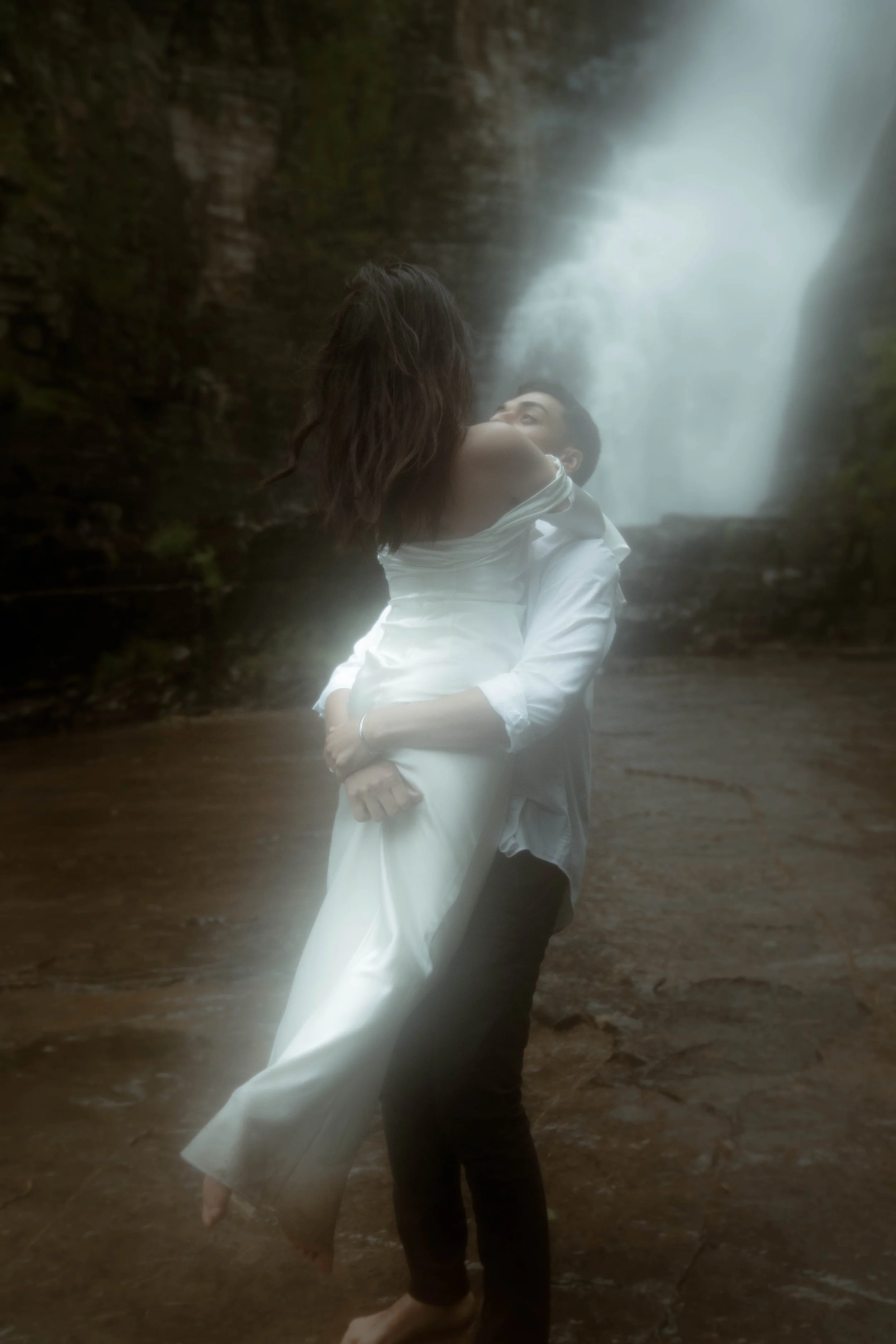 Un couple danse devant une cascade, la femme porte une robe blanche et l'homme une chemise blanche. La scène est en extérieur, humide avec de l'eau et de la pierre en arrière-plan.