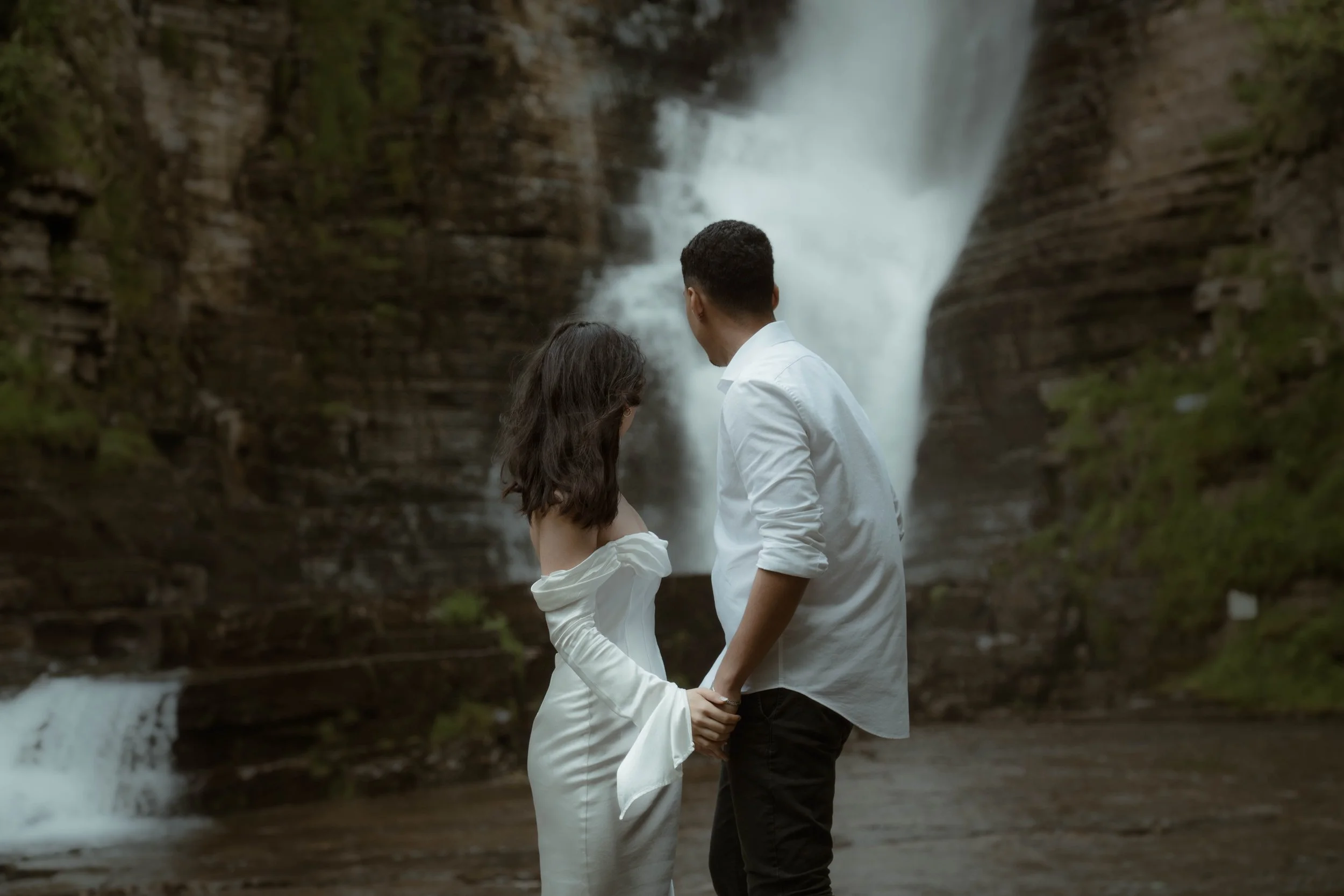 Un couple habillé en blanc se tient près d'une cascade dans la nature.