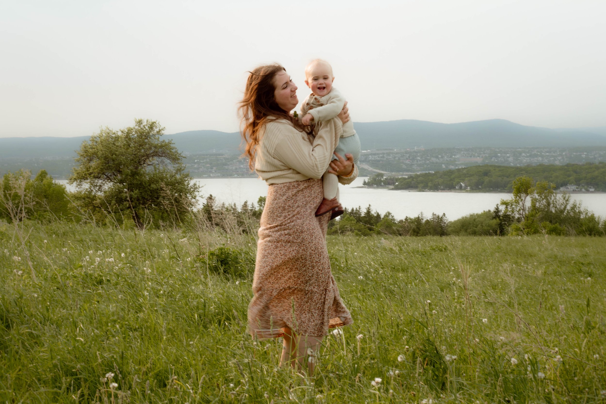 Une femme souriante tenant un bébé dans un paysage naturel avec des arbres, une rivière et des montagnes en arrière-plan.