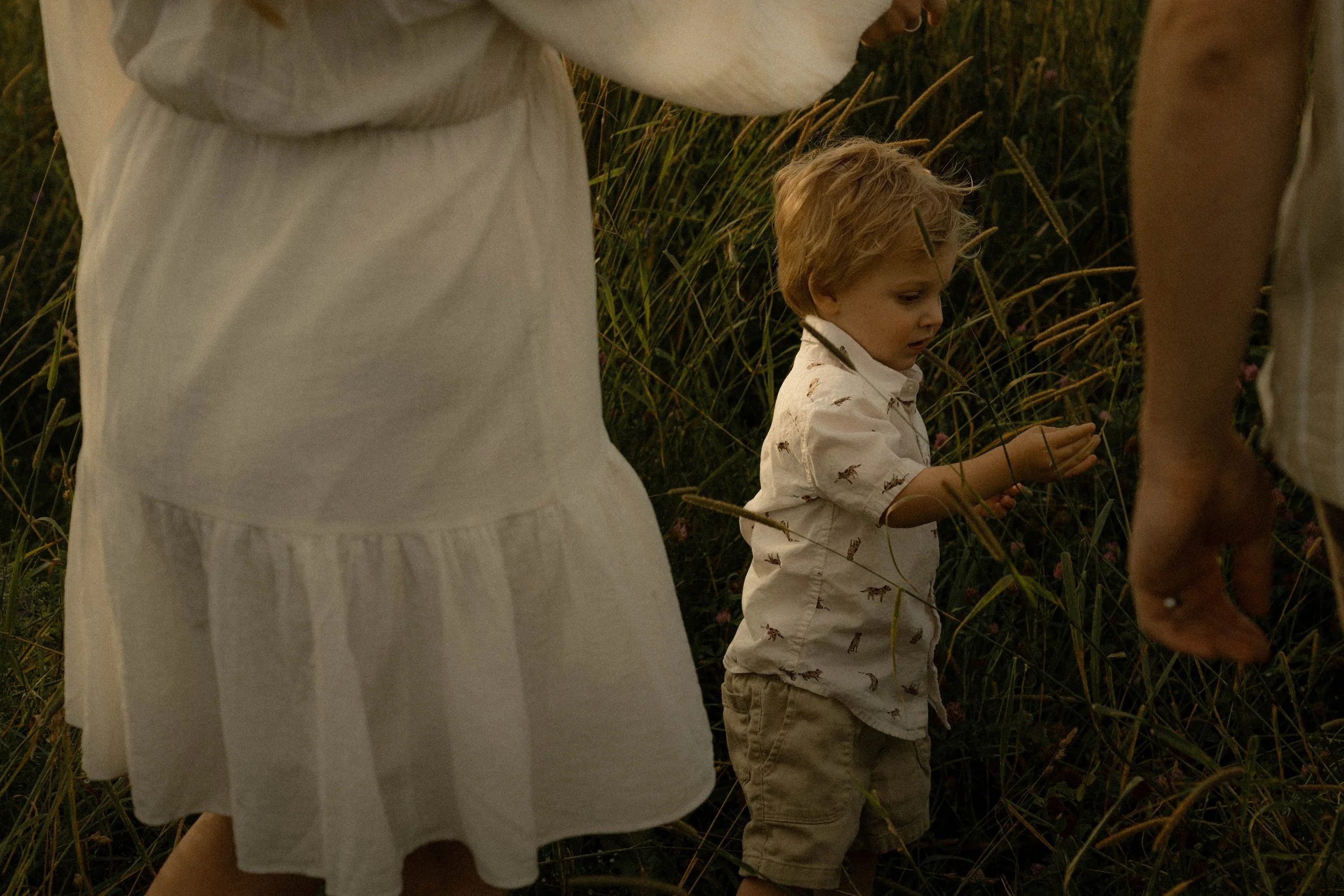Deux enfants en chemise claire, parmi de hautes herbes, probablement dans un champ ou une prairie, le soir ou au crépuscule.