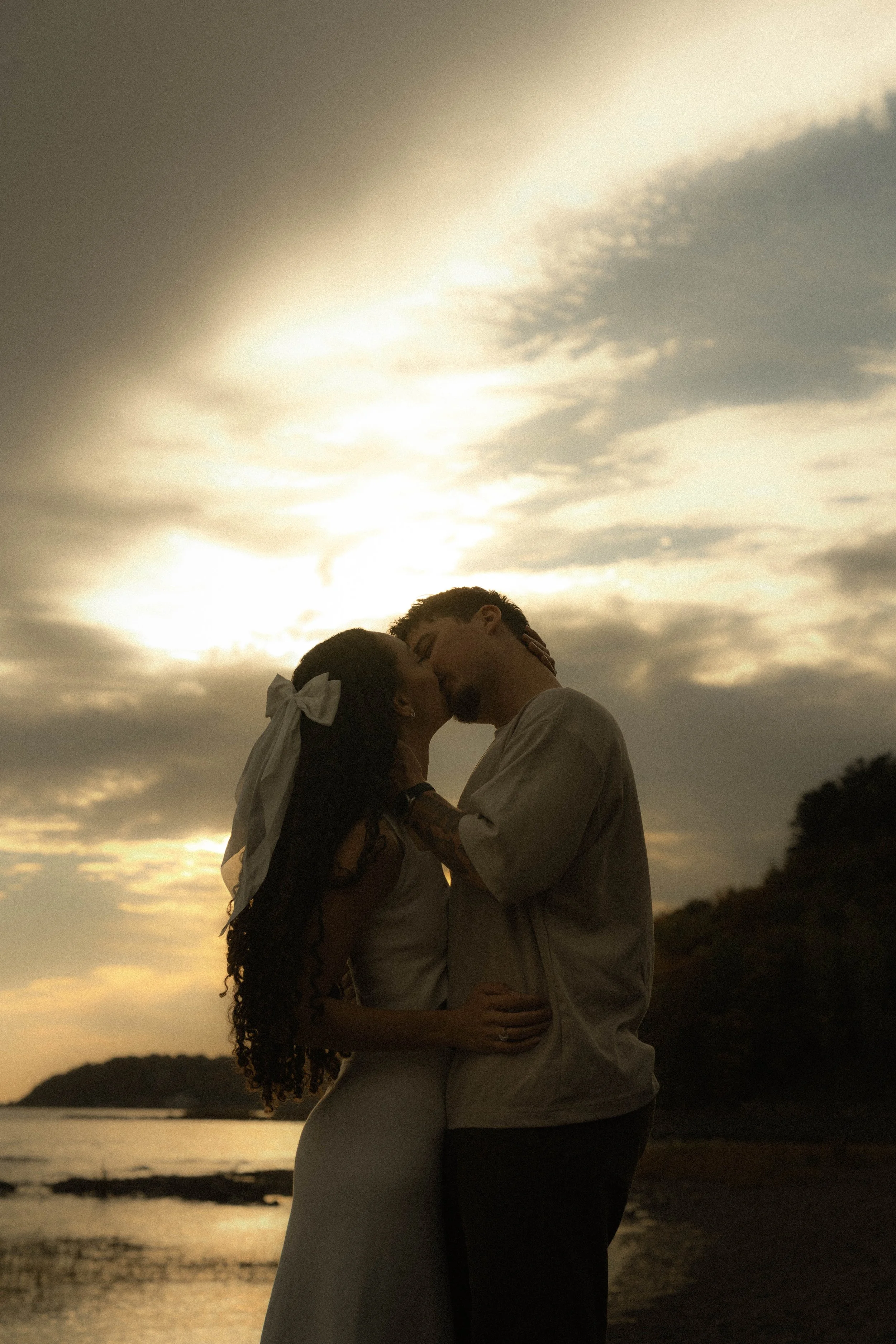 Un couple s'embrassant tendrement lors d'un coucher de soleil sur la plage, avec une mer calme en arrière-plan et un ciel nuageux aux teintes dorées.