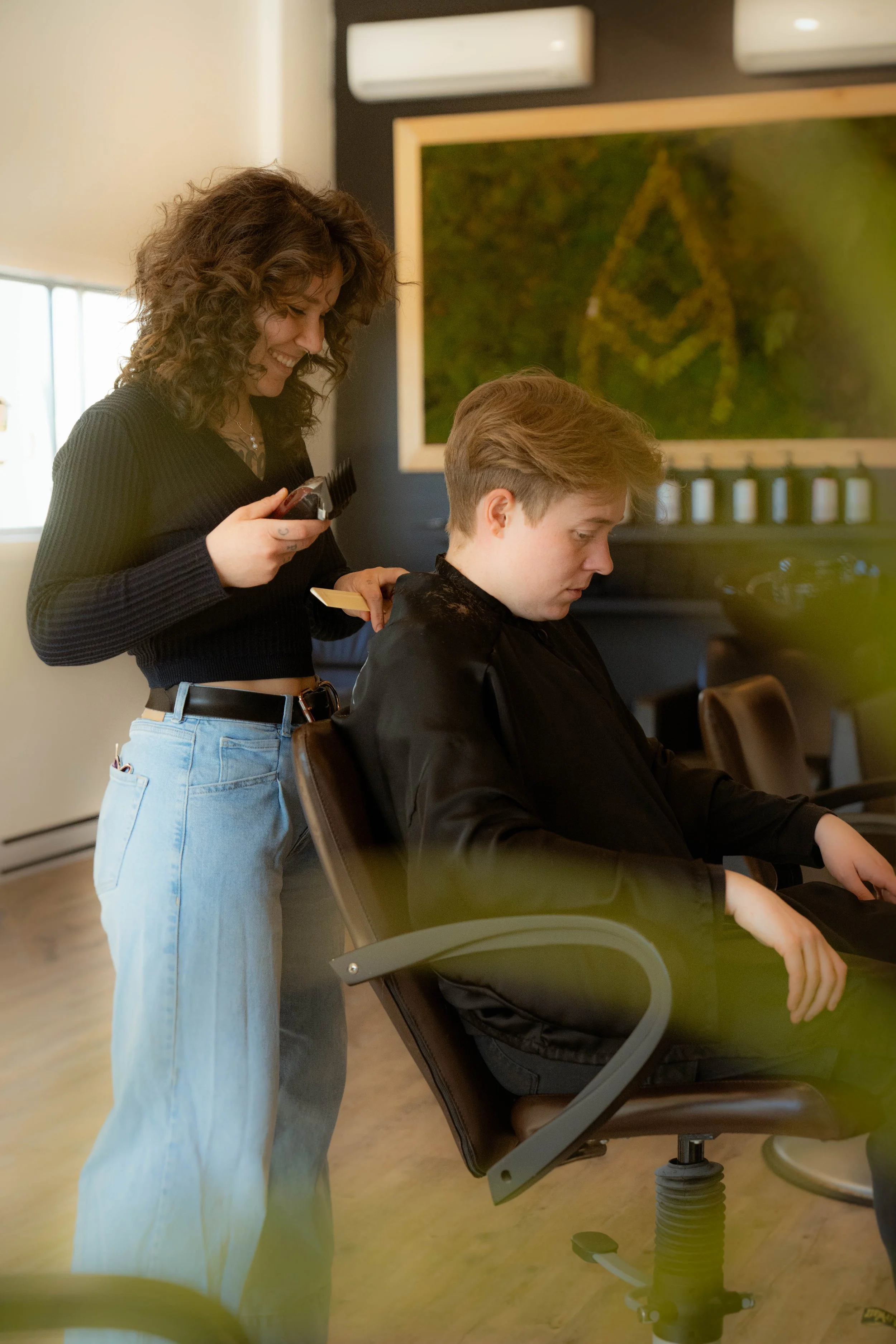 Une femme coiffant un jeune homme assis dans un salon de coiffure, avec une peinture verte en arrière-plan.
