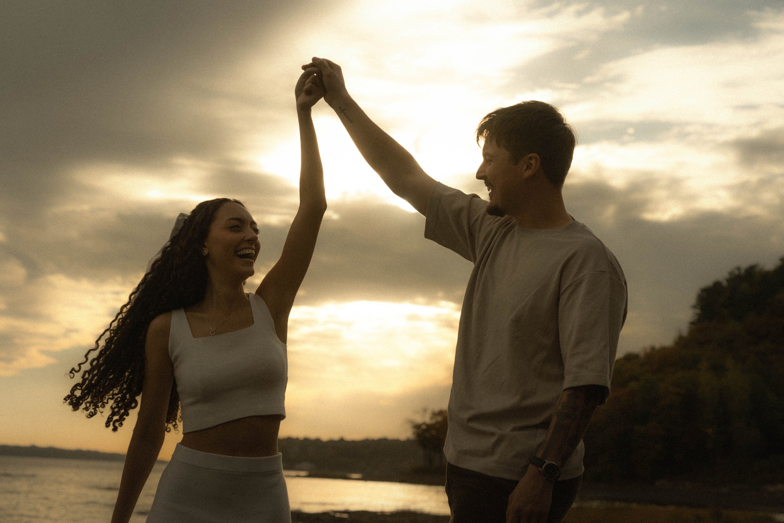 Un couple souriant lors d'un coucher de soleil au bord d'une rivière, se tenant par la main en hauteur.