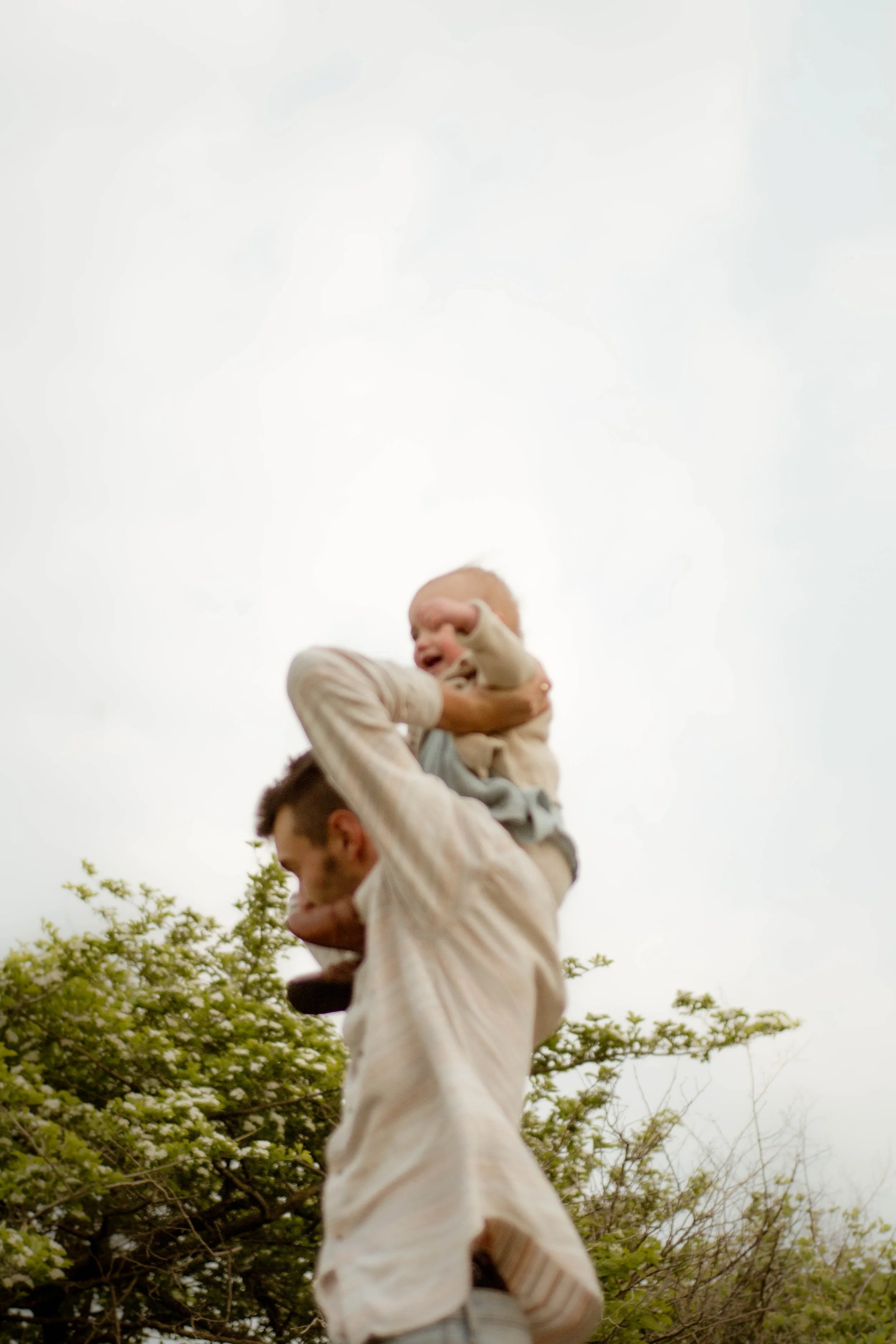 Un enfant assis sur les épaules d'un adulte, riant avec les bras levés, entouré d'arbres en fleurs sous un ciel nuageux.
