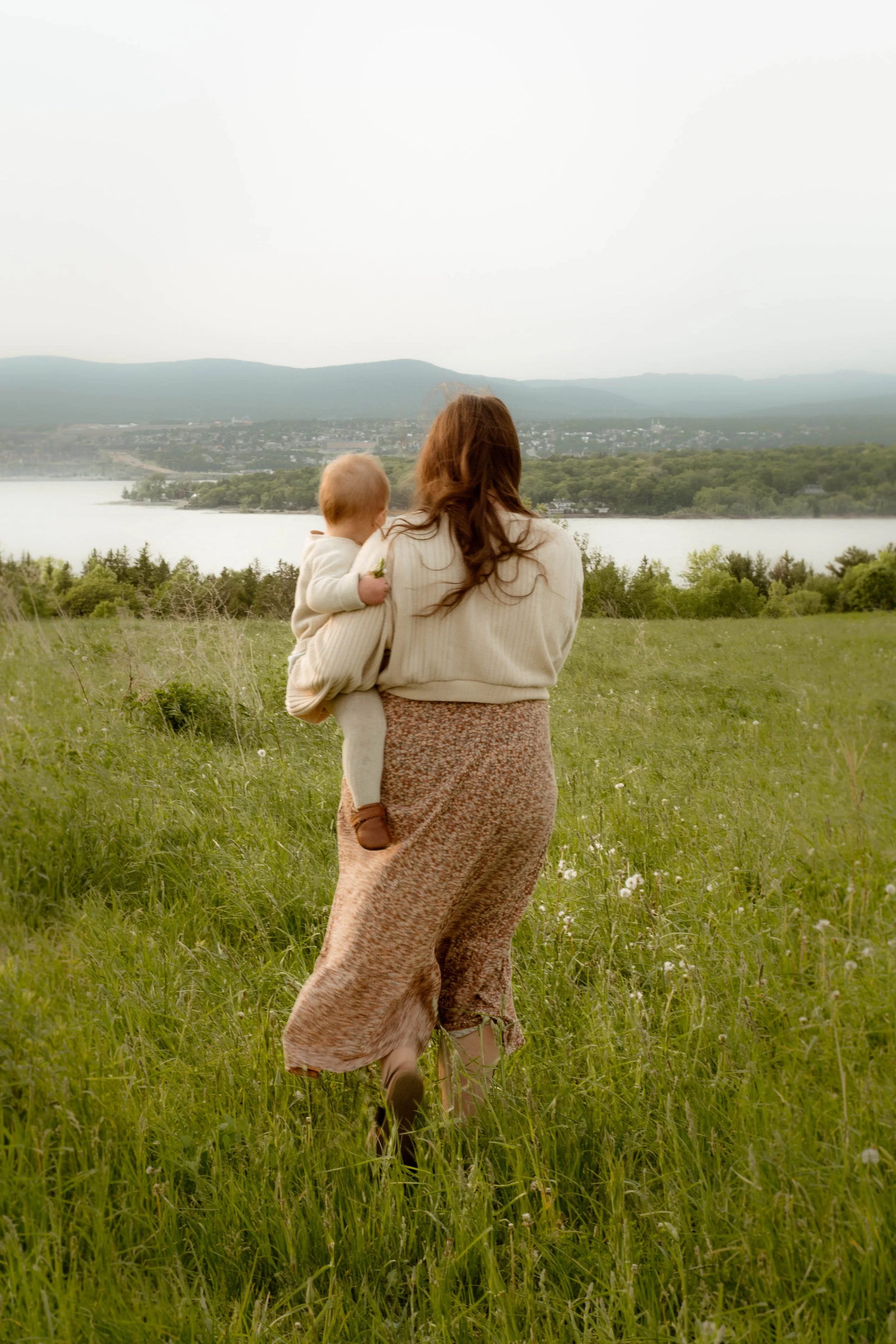 Une femme portant une robe longue marche dans un champ d’herbe verte avec un enfant dans ses bras, regardant vers un lac et des collines au loin, par une journée nuageuse.