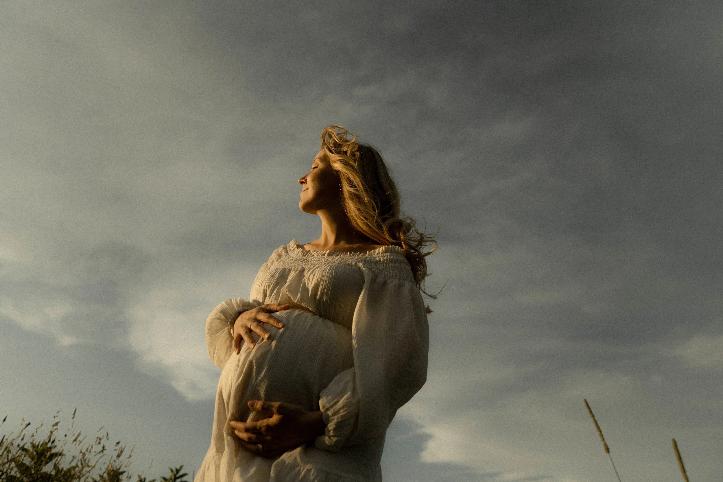 Femme blonde en vêtement beige, bras croisés, regardant vers la gauche, sous un ciel nuageux, dans un champ près de végétation.