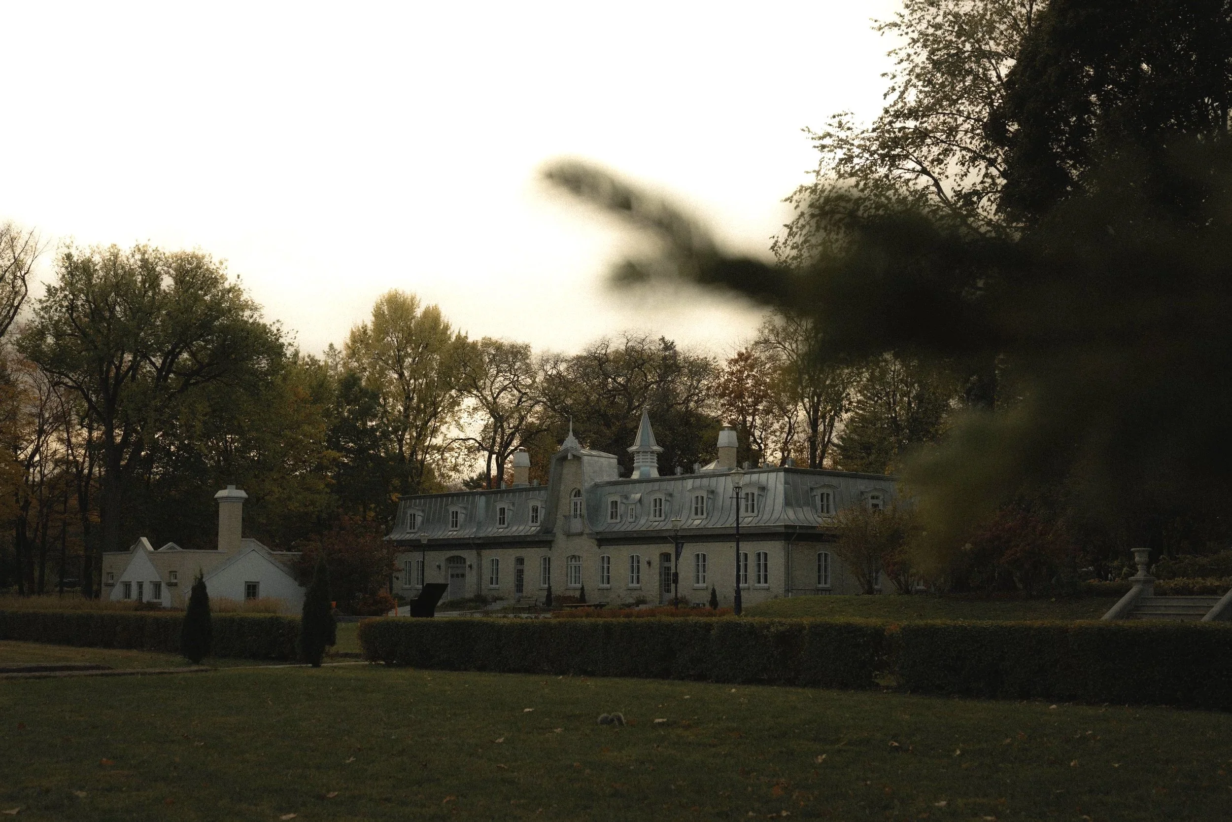 Un grand bâtiment blanc avec des toits en formes pointues, entouré d'arbres avec des feuilles orange, jaunes et vertes. L'image est sombre et semble être prise au crépuscule ou par temps nuageux.