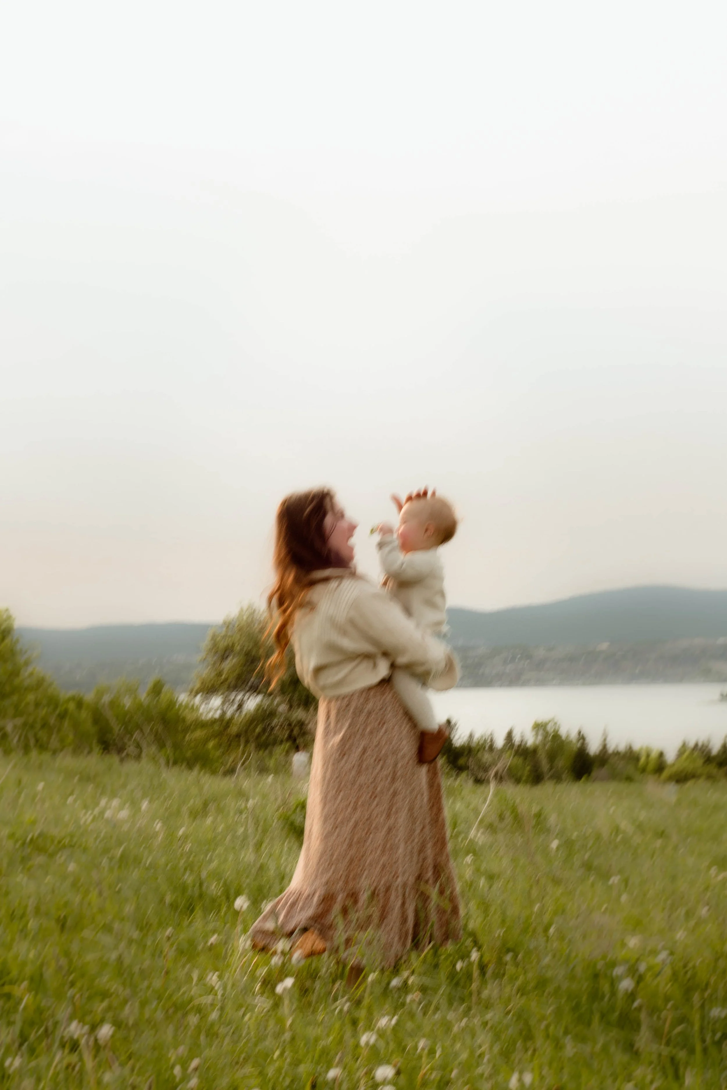 Une femme tenant un enfant dans un paysage naturel avec un lac et des montagnes en arrière-plan. Le ciel est clair et la scène est paisible.
