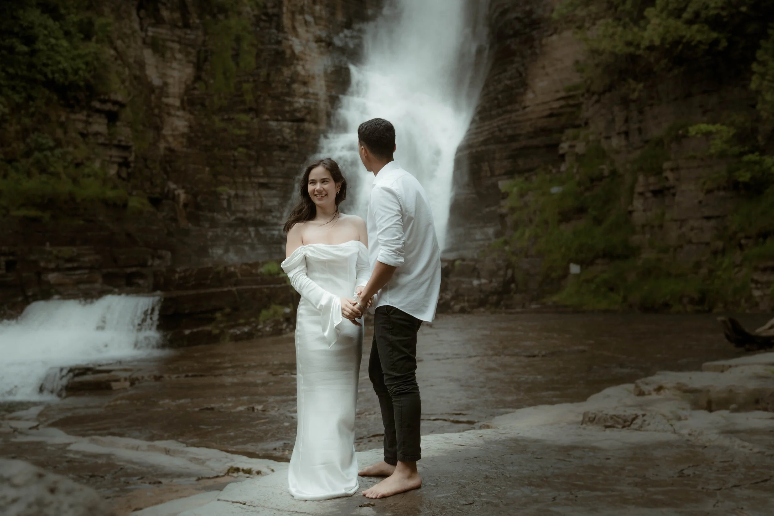 Un couple devant une cascade, la femme en robe blanche et l'homme en chemise blanche et pantalon noir, se tenant la main sur une roche près de l'eau.