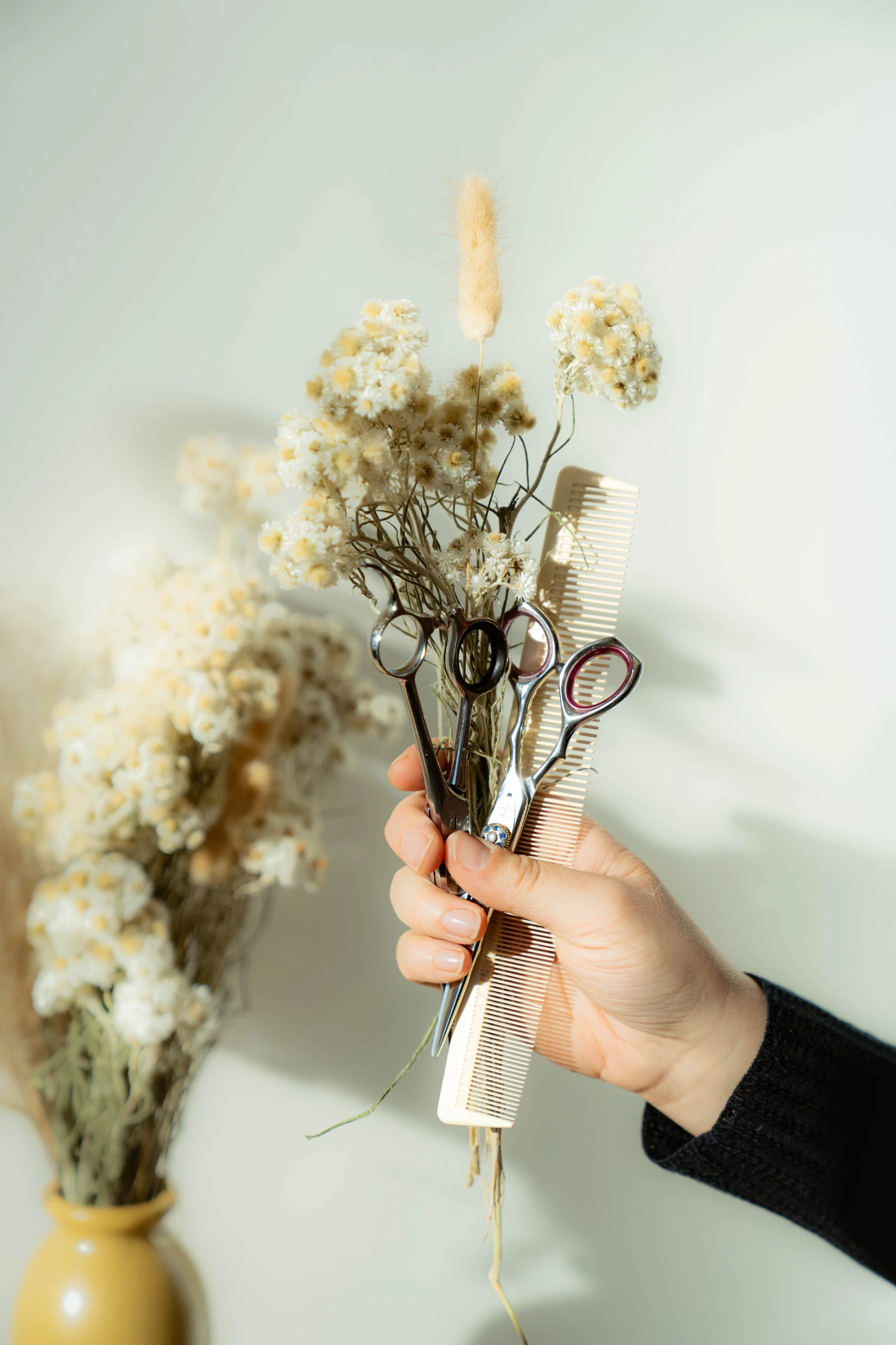 Une main tenant une paire de ciseaux, un peigne et des fleurs séchées dans un vase en arrière-plan, dans une ambiance claire et minimaliste.