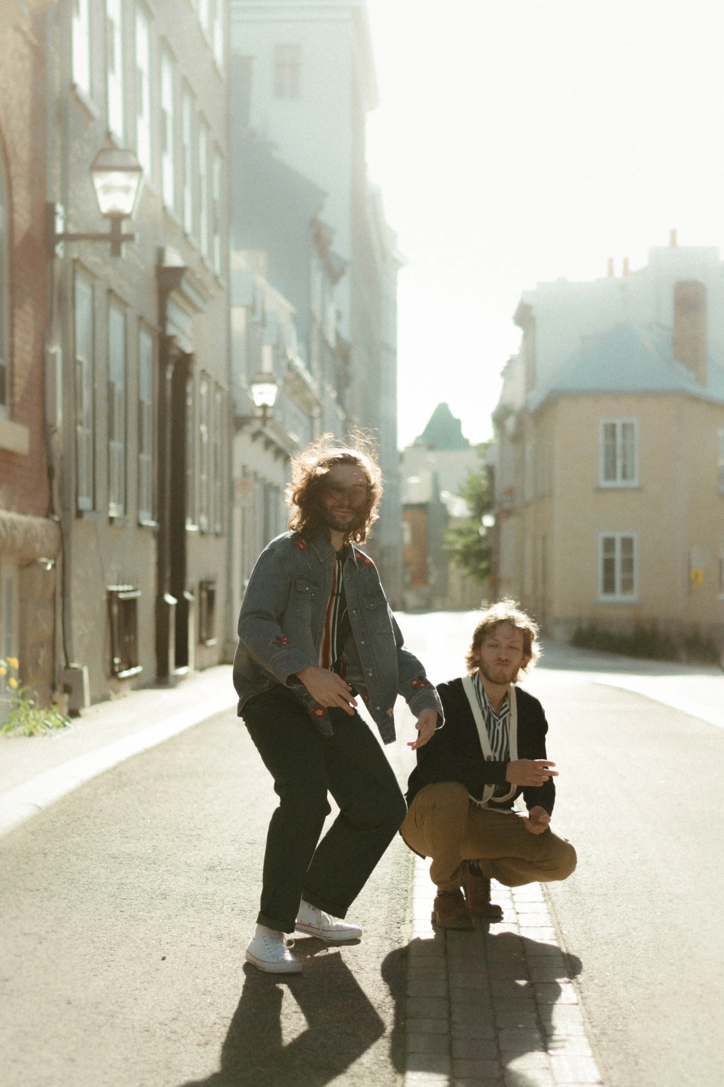 Deux jeunes hommes posent dans une rue lumineuse, l'un en position accroupie, l'autre debout, avec des bâtiments résidentiels en arrière-plan et la lumière du soleil créant des ombres marquées.