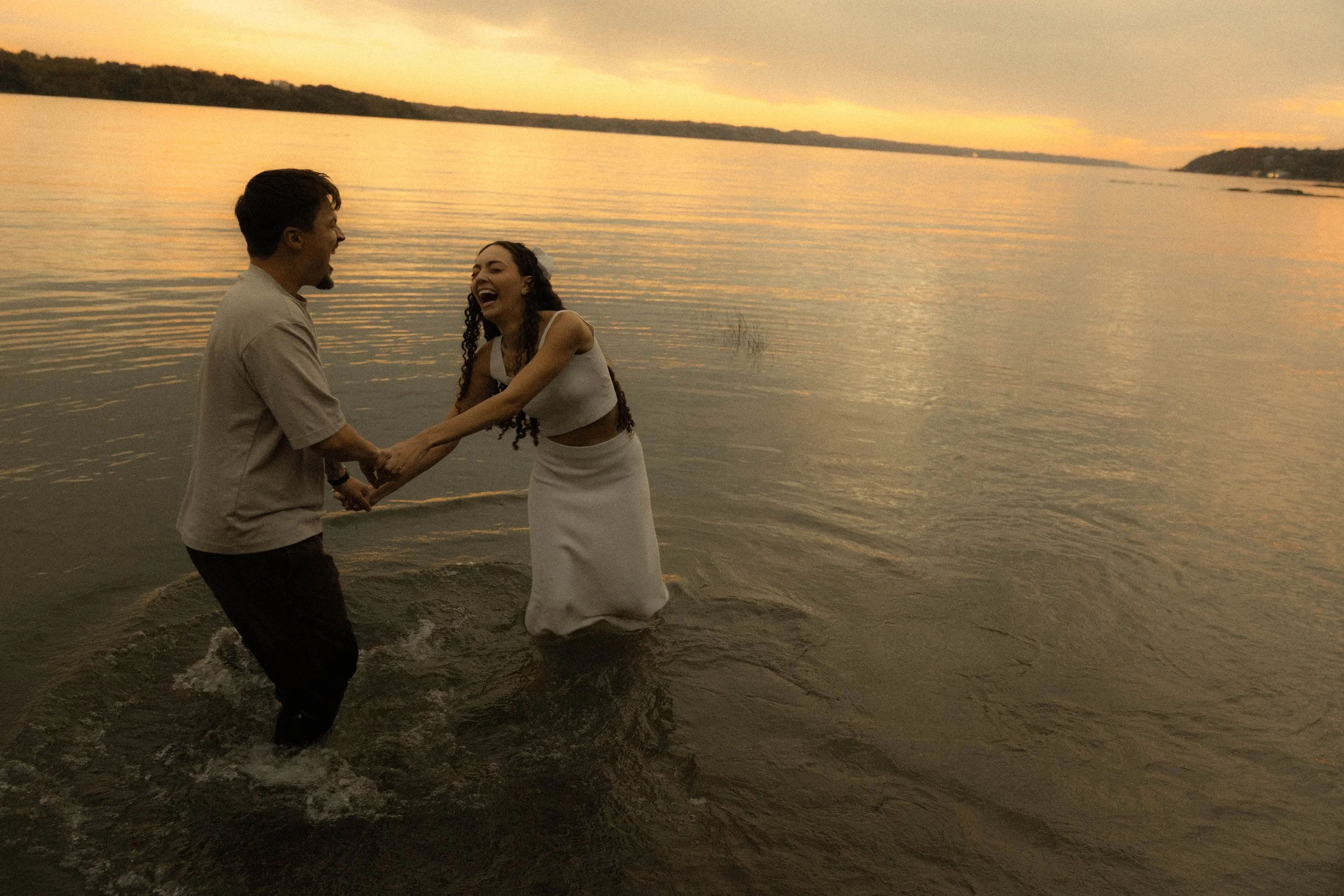 Un couple en robe blanche jouent ensemble dans l'eau au bord d'un lac au coucher du soleil, riant et tenant la main, avec un paysage de collines en arrière-plan.
