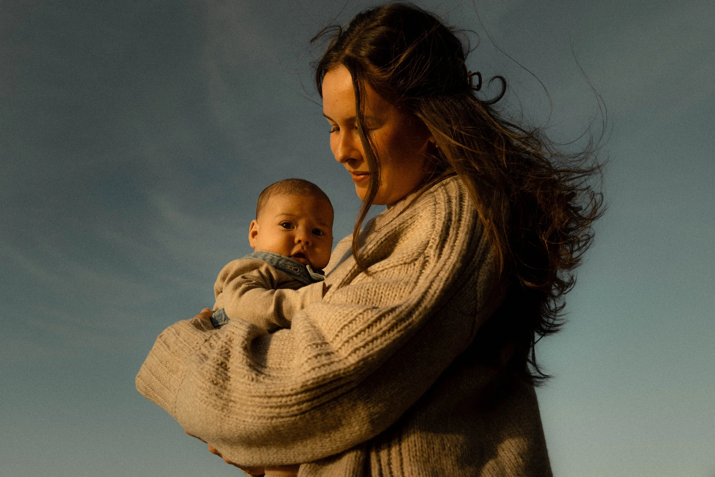 Une femme portant un manteau beige tient un bébé dans ses bras. La mère regarde vers le bas, tandis que le bébé regarde vers l'avant, sous un ciel dégagé.
