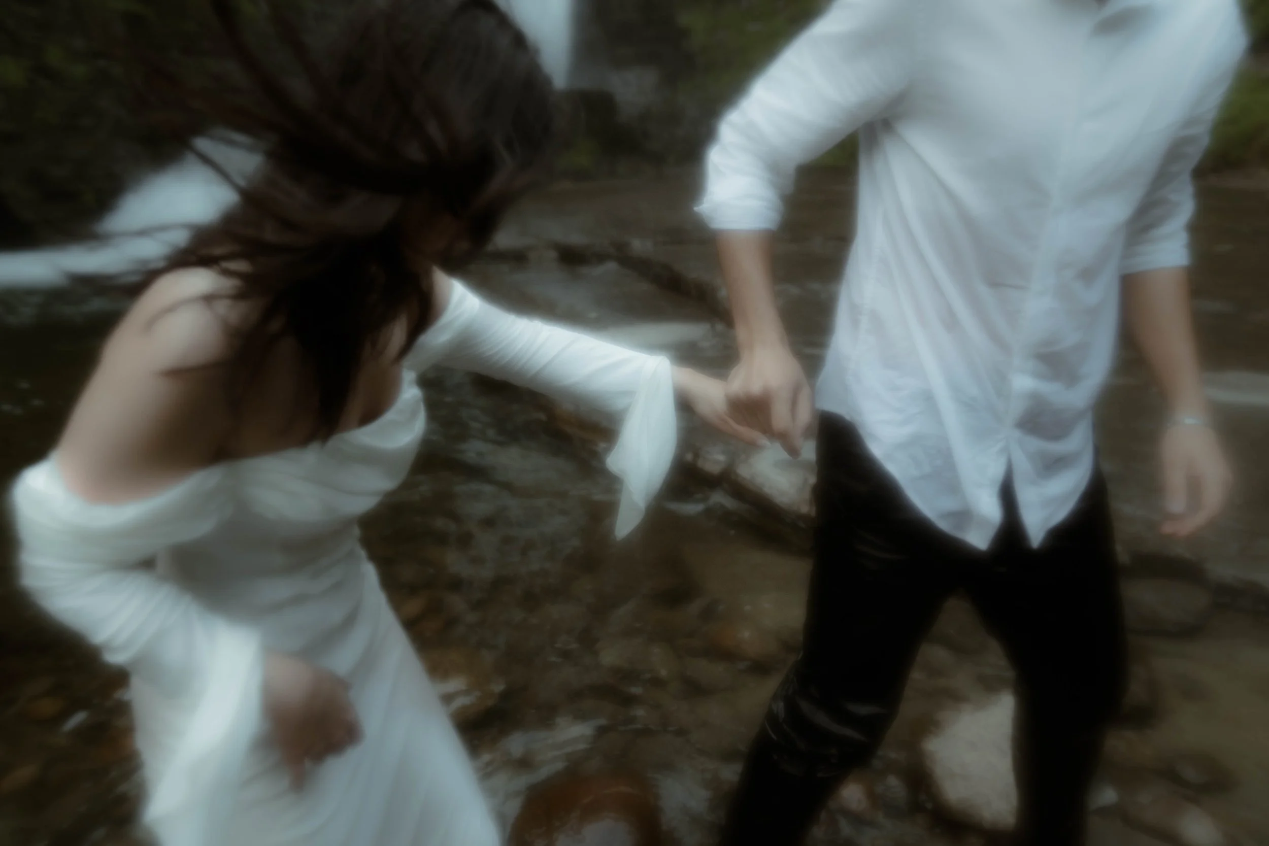 Deux personnes, l'une en robe blanche et l'autre en chemise blanche, se tiennent par la main dans une rivière. La personne en chemise a le bras tendu vers la personne en robe. La scène est naturelle avec de l'eau courante et un environnement forestie