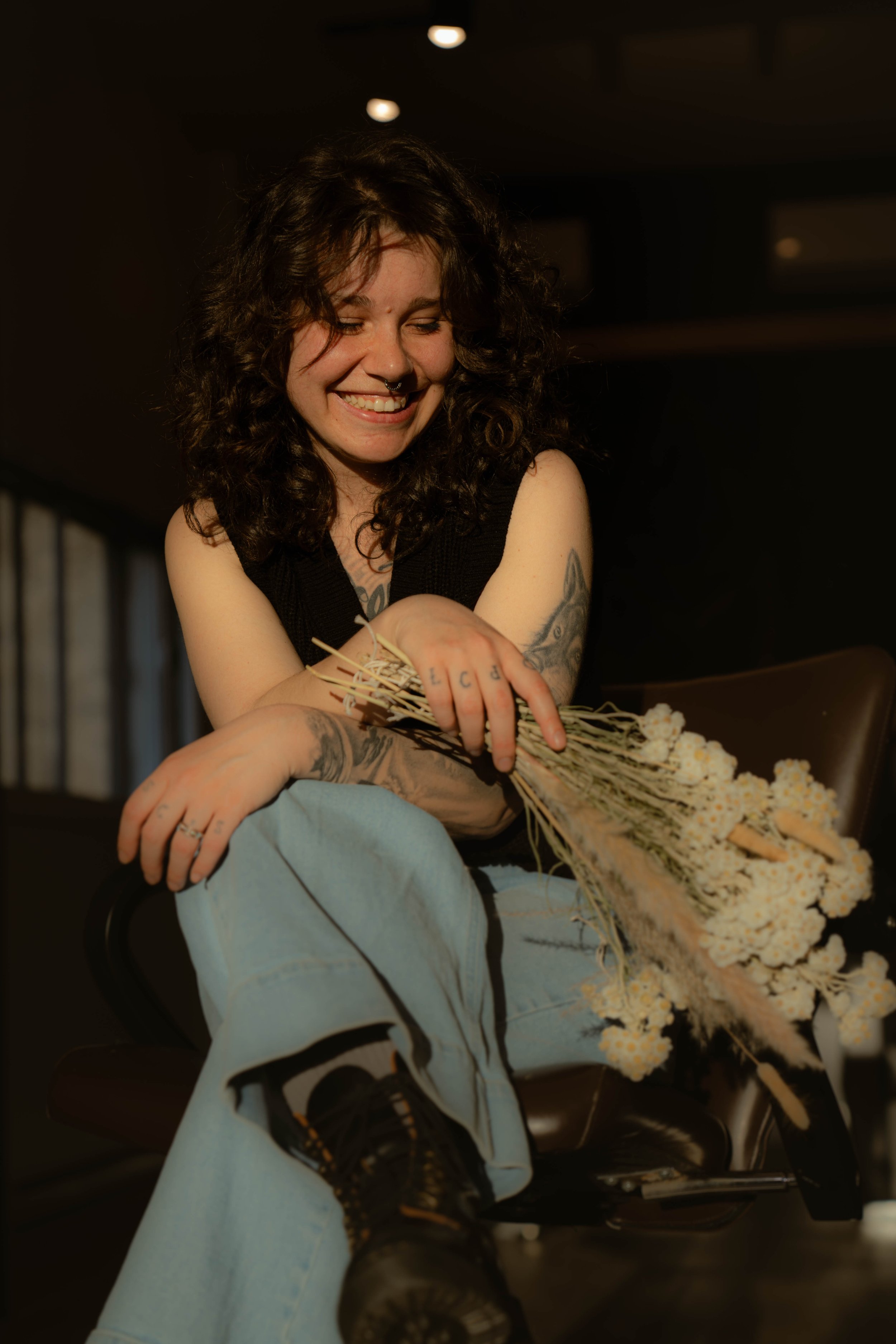 Une jeune femme aux cheveux bouclés, souriante, portant un tatouage sur le bras, en train de tenir un bouquet de fleurs blanches.