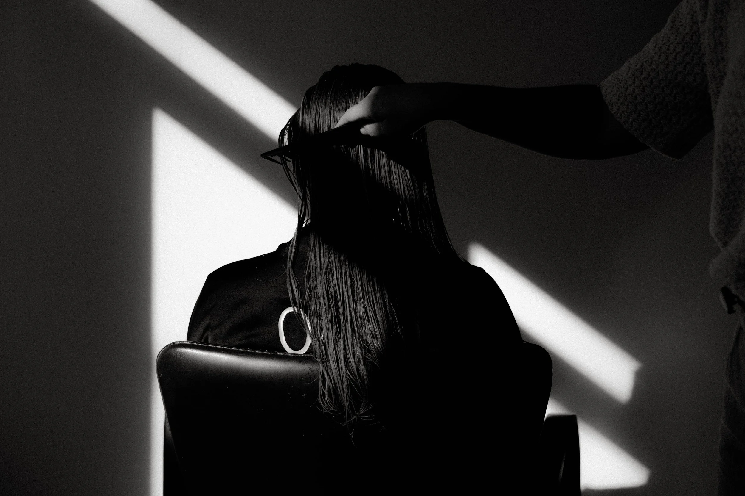 Une personne coiffée d'une tignasse longue et mouillée est assise, portant un t-shirt noir. Une main de quelqu'un d'autre, portant un pull en laine, ajuste ou lave ses cheveux, dans un environnement sombre avec des rayons de lumière diagonaux.
