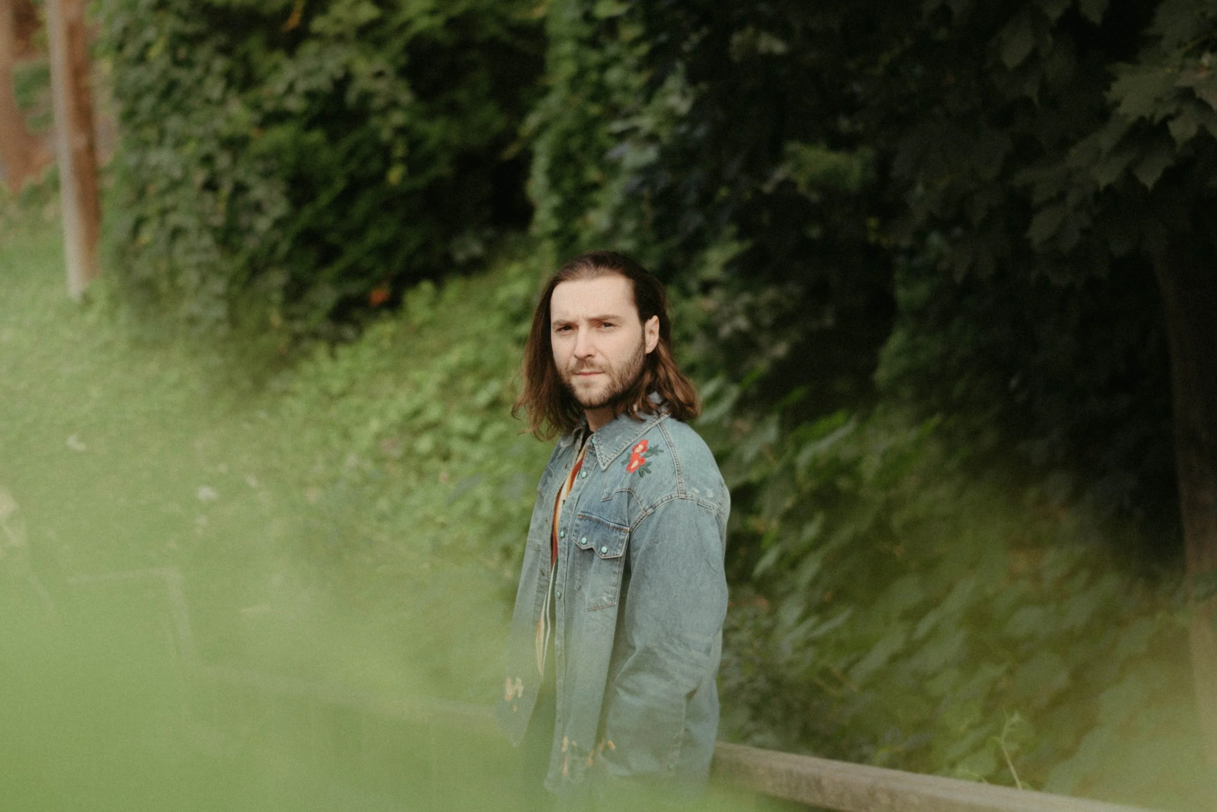 Un homme à cheveux longs et barbe porte une veste en jean avec des broderies rouges, il se tient dans un espace vert entouré d'arbres et d'arbustes.