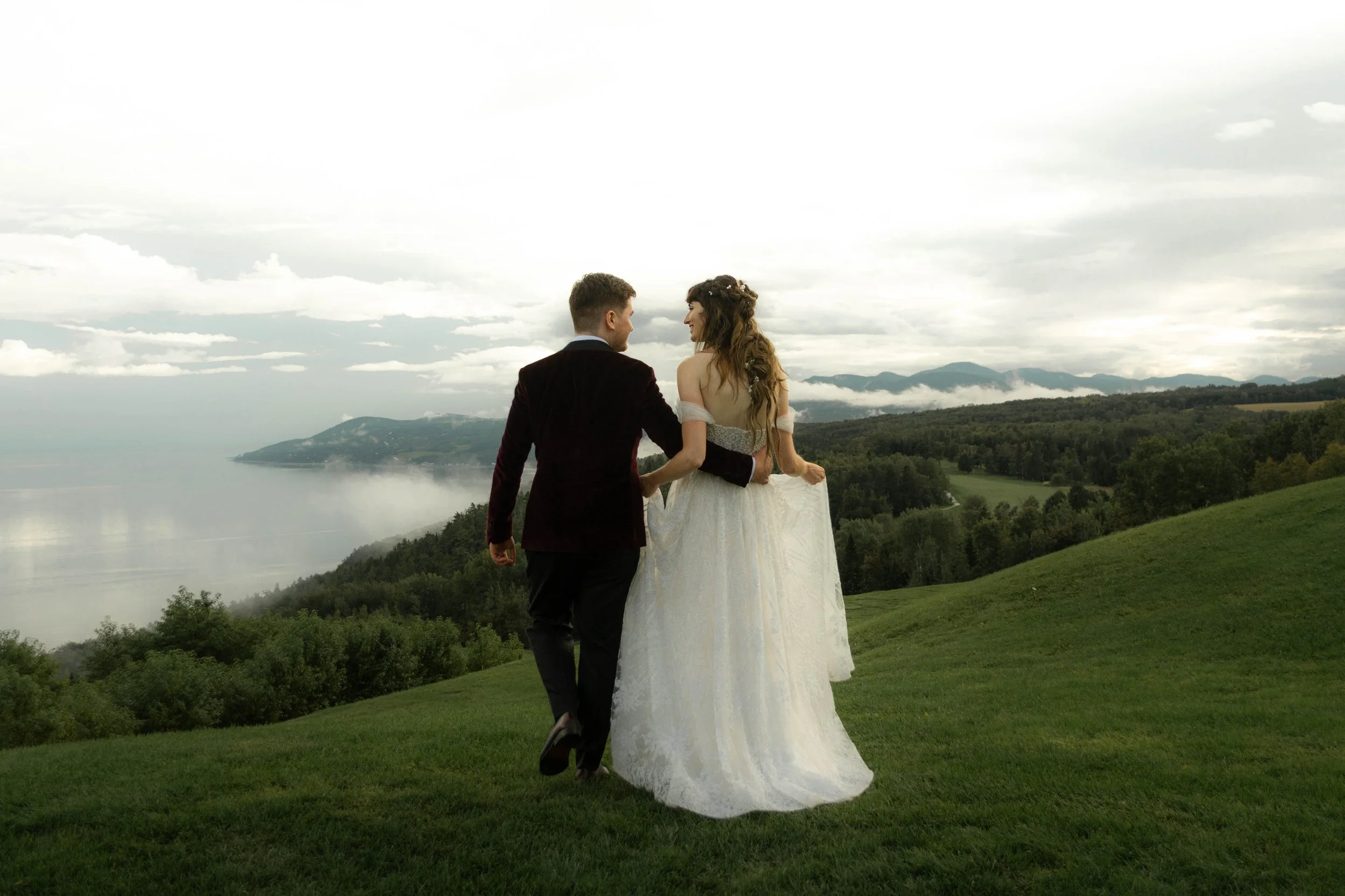 Un couple en mariage marchant dans un paysage verdoyant avec des montagnes au loin et un ciel nuageux