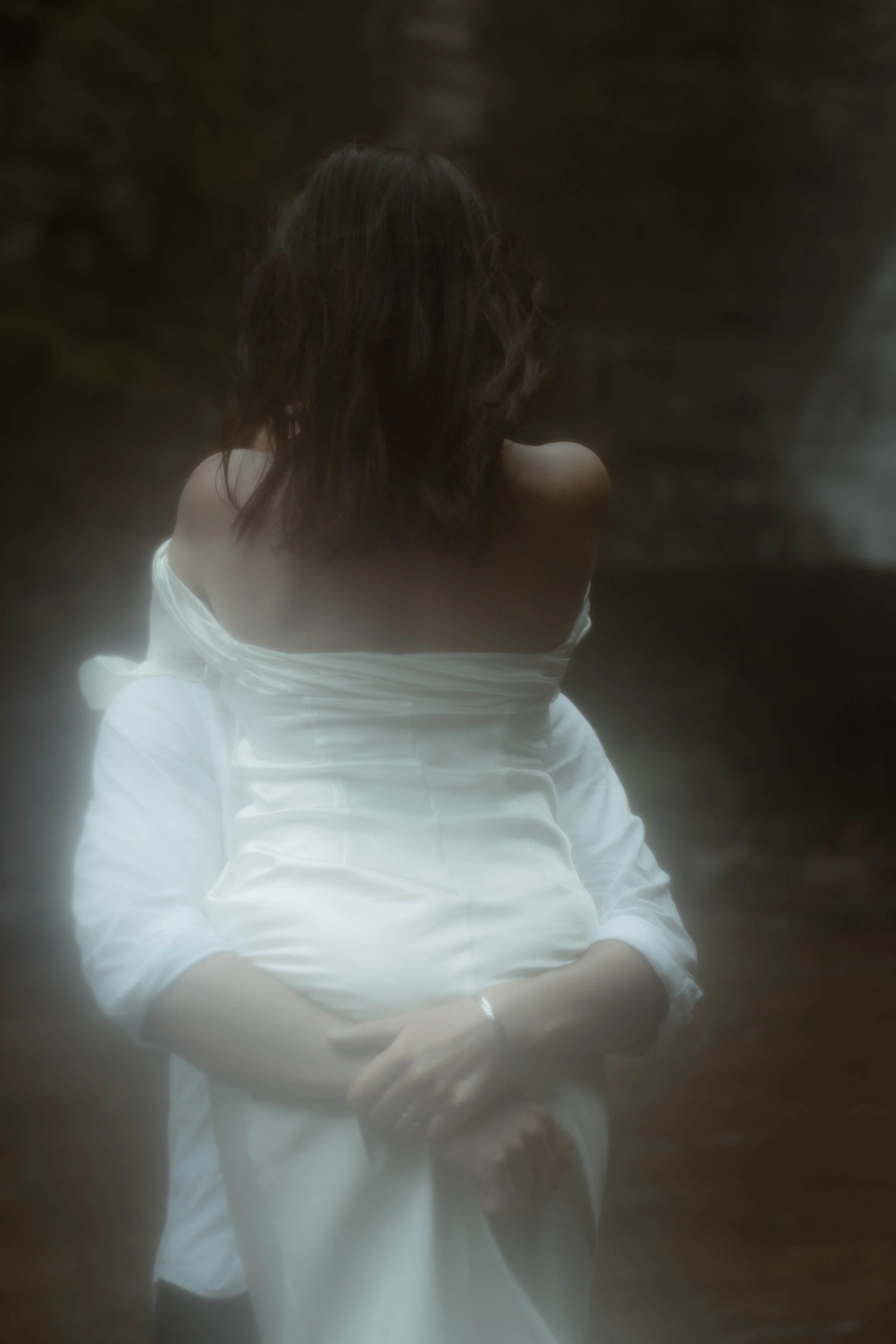 Femme vue de dos, portant une robe blanche, cheveux longs et ondulés, dans un environnement sombre.