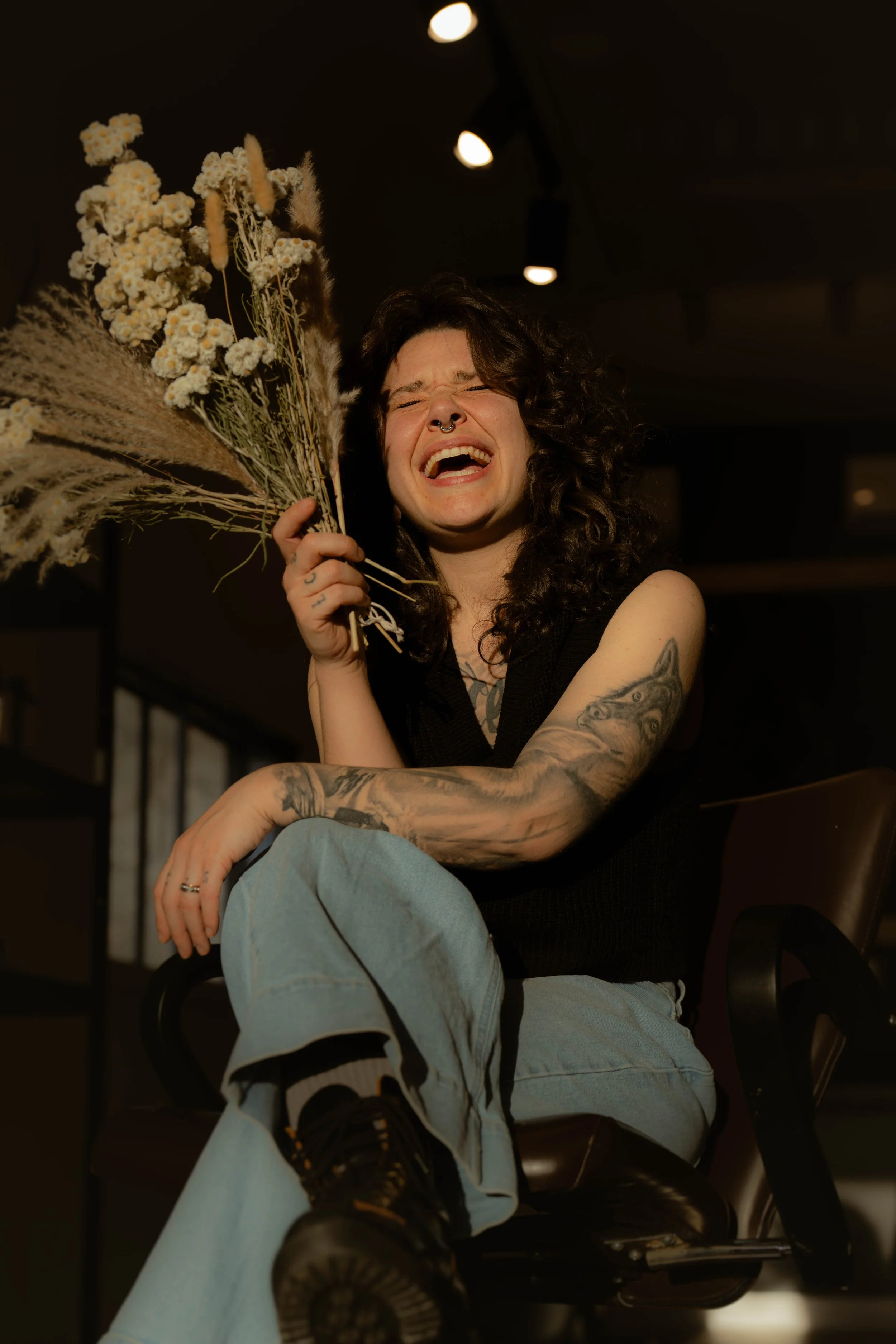 Une femme avec des tatouages, des boucles d'oreilles et un piercing, riant et tenant un bouquet de fleurs séchées, assise dans un espace intérieur sombre.