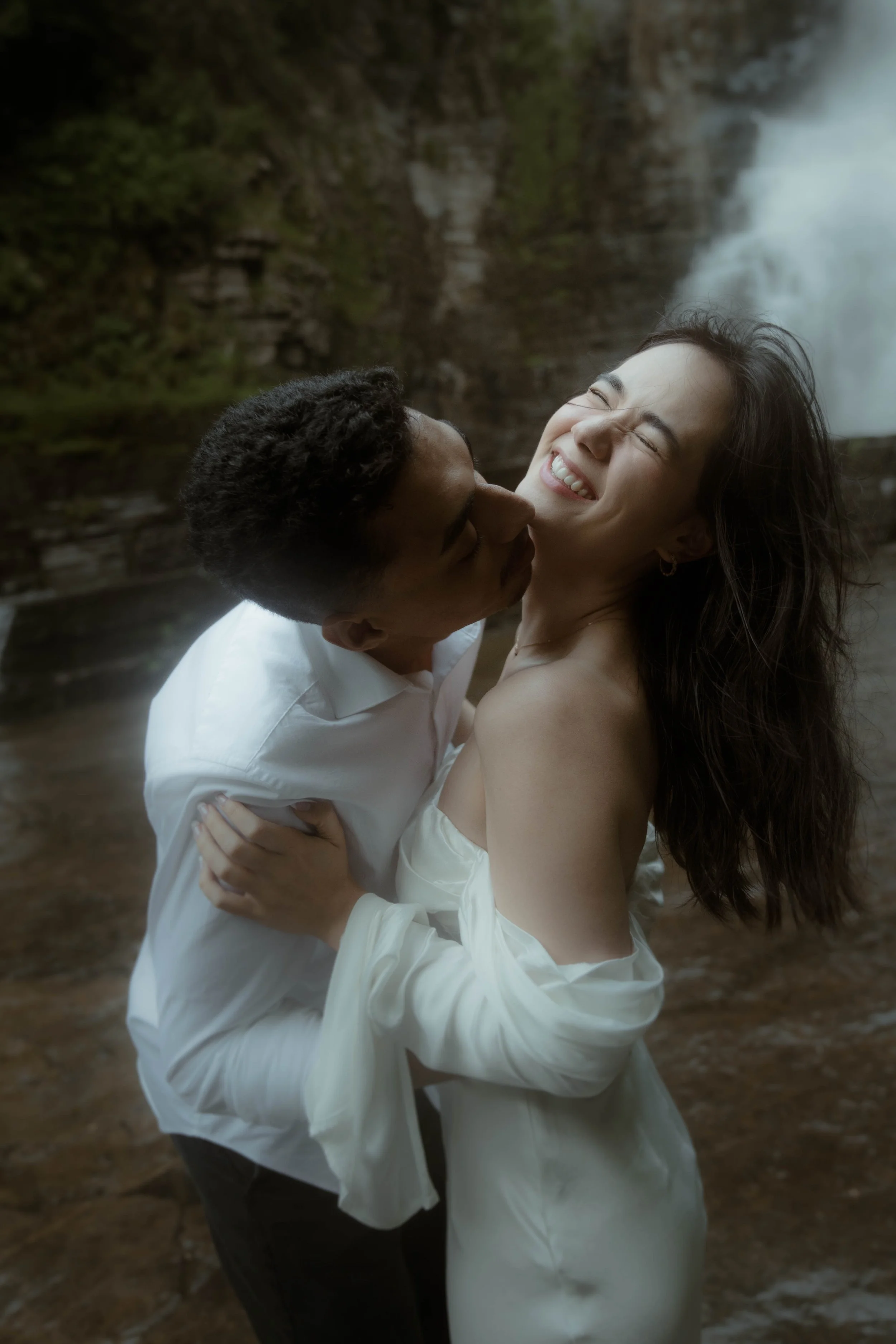 Un couple souriant et en train de s'embrasser près d'une cascade. L'homme porte une chemise blanche, la femme porte une robe blanche.