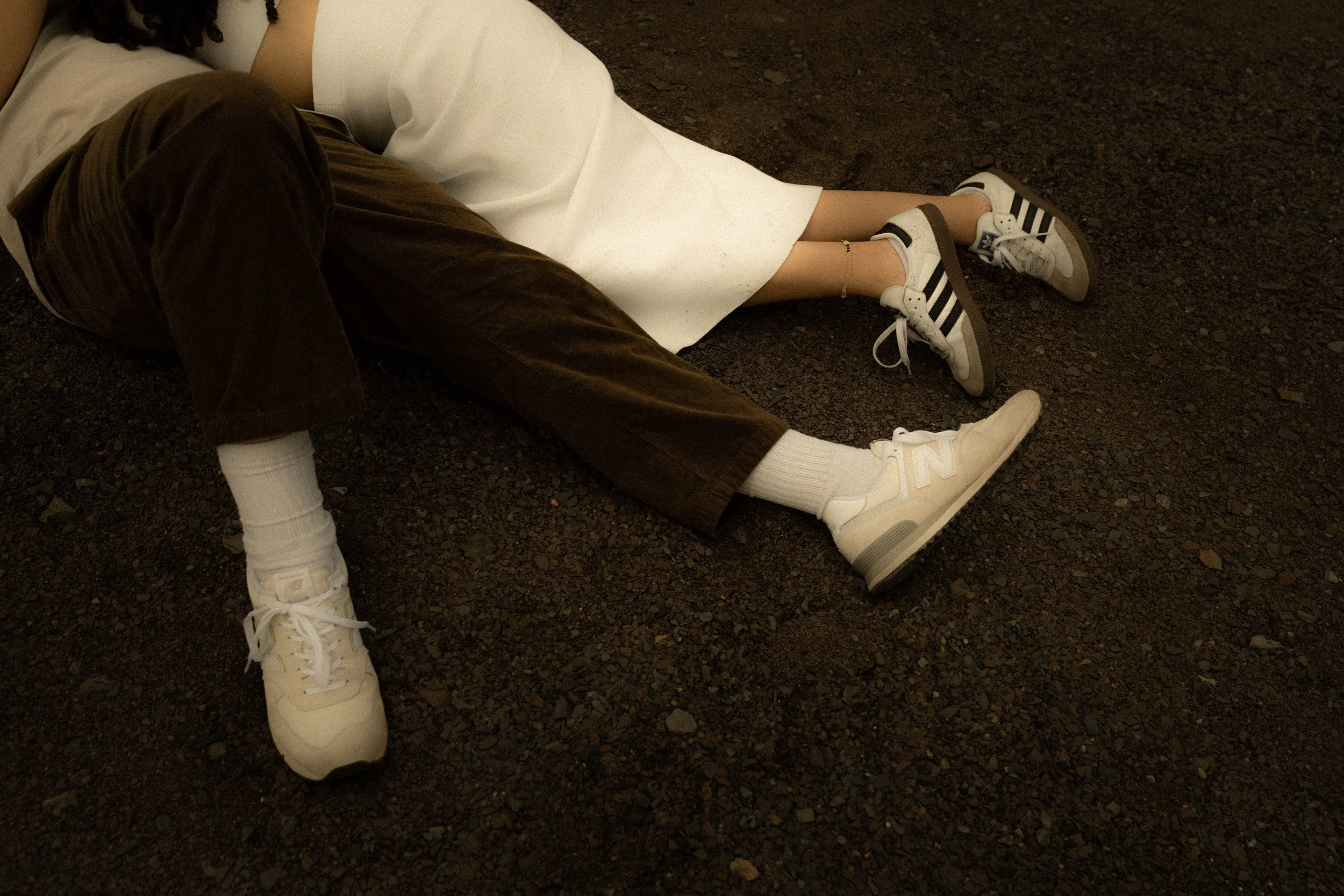 Deux personnes sont allongées sur le sol en pierre ou en gravier, en position de repos ou de repos. Une personne porte des chaussures de sport blanches et noires, une autre porte des chaussures blanches.
