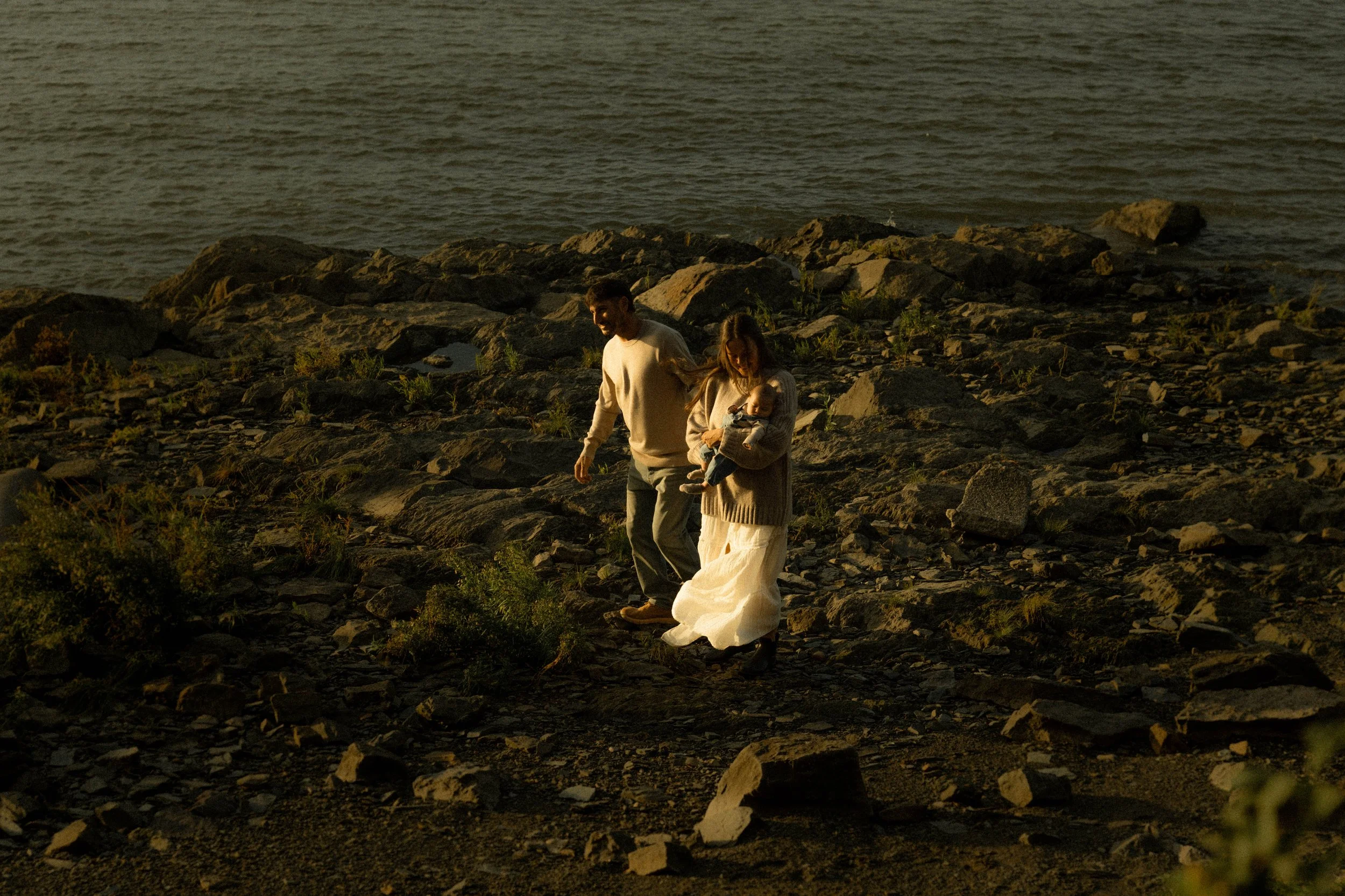 Un couple avec un bébé marche sur des rochers près de l'eau au coucher du soleil.