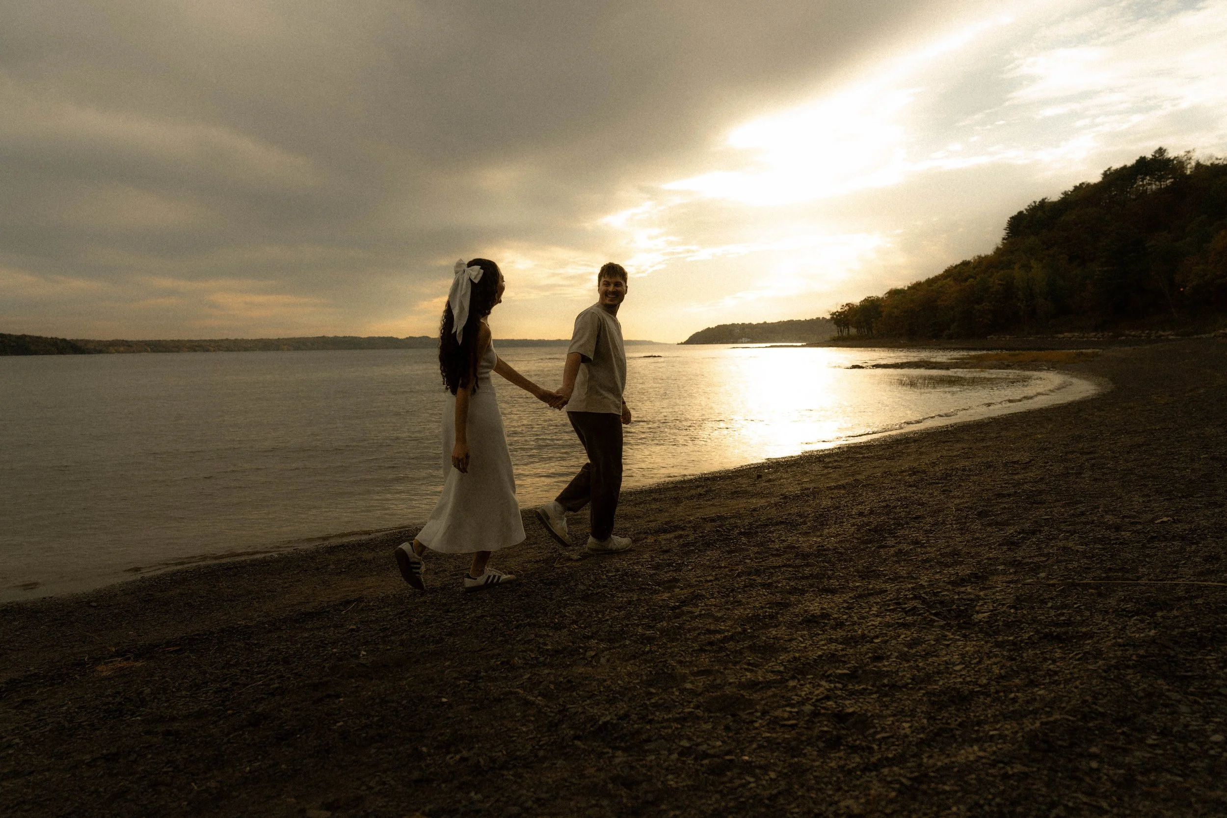 Un couple marche main dans la main sur une plage au coucher du soleil, avec un ciel partiellement nuageux et des arbres en arrière-plan.