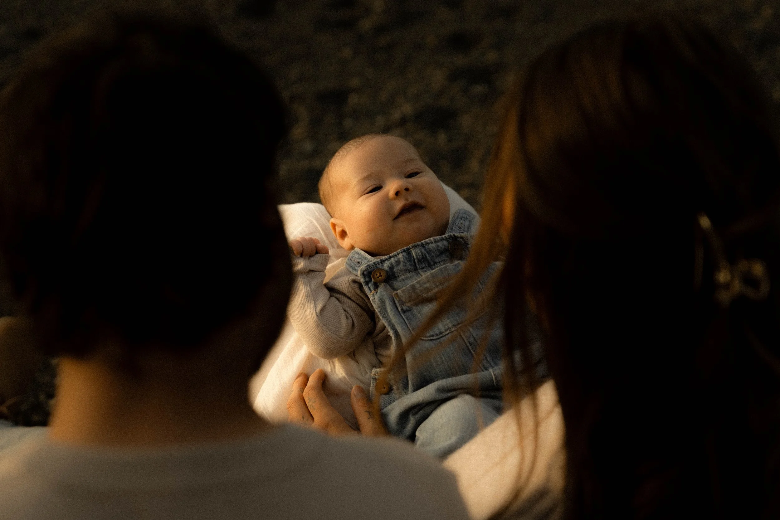 Un bébé sourit, vu à travers les cheveux et les épaules de deux adultes qui l'entourent, dans un environnement intérieur chaleureux.